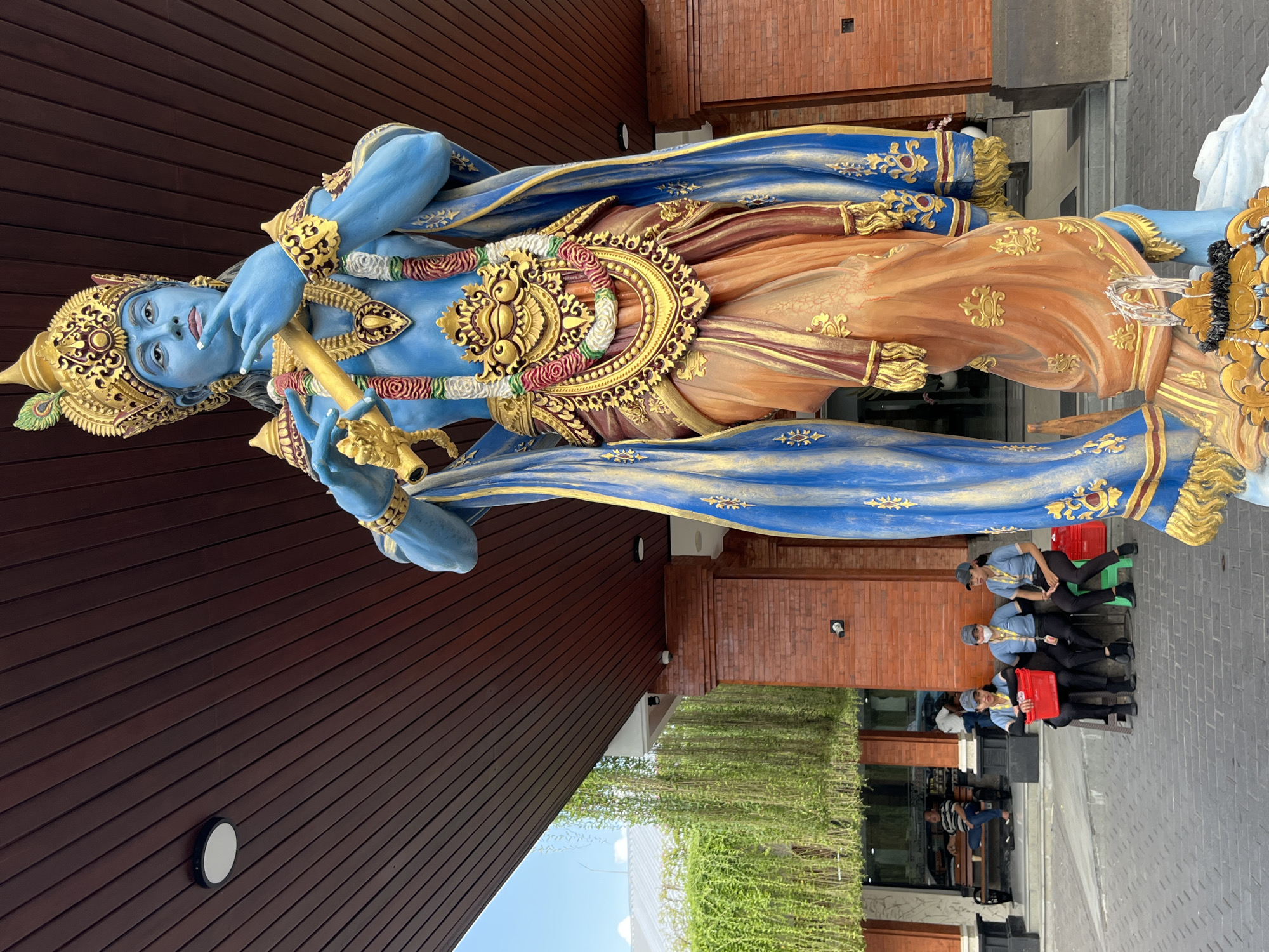 A towering blue statue of a deity, likely Krishna, stands prominently in an outdoor setting. The statue is adorned with elaborate gold jewelry and ornate clothing in shades of blue and orange, with a gold headdress. It holds a golden staff. To the right of the statue, a group of people are seated on benches and the ground. Some are wearing blue shirts, dark pants, and caps, and appear to be taking a break or waiting. A few red containers are visible among them. In the background, there is a brick wall, a building with wooden paneling, and lush green foliage cascading down a structure. The ground is paved with interlocking bricks. The lighting suggests it is daytime, with bright sunlight casting shadows. The overall scene appears to be a public space, possibly a temple or a cultural attraction in Kuta, Indonesia. There is no visible text.