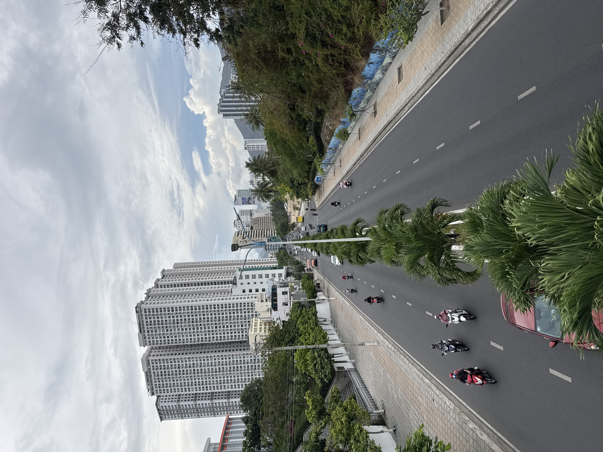 Captured from an elevated position, the image depicts a straight, multi-lane road in Bac Nha Trang Ward, Vietnam, flanked by high-rise buildings on one side and a lower-level sidewalk and vegetation on the other. A line of palm trees runs along the median of the road. Multiple motorbikes are moving along the road. A red car is partially visible in the foreground. The sky is cloudy. The architectural styles of the buildings on the left are modern and high-density.