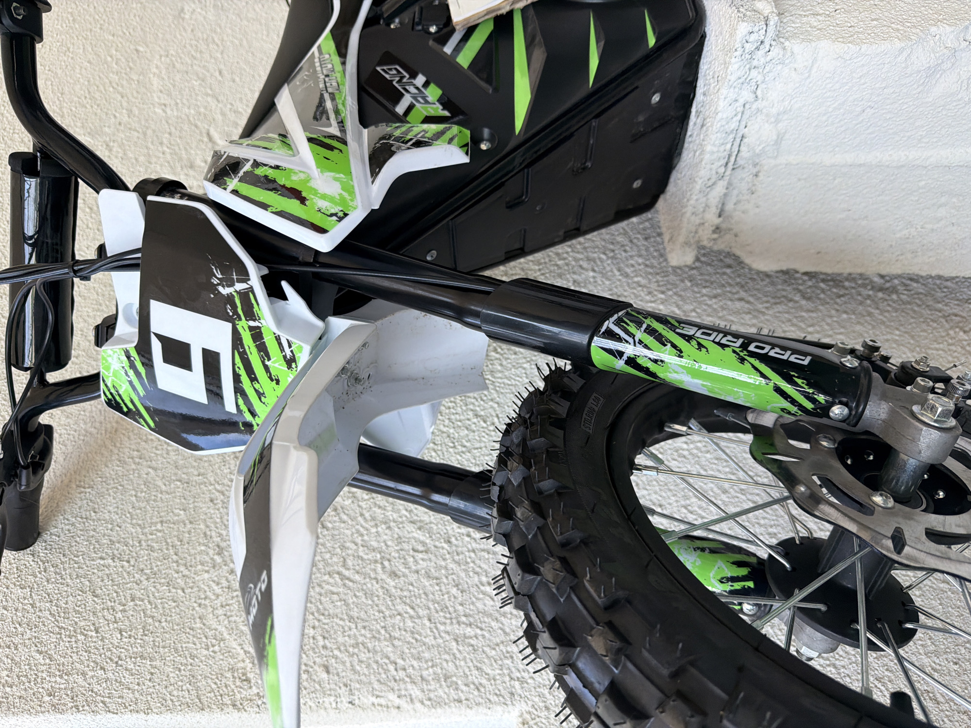 A close-up, eye-level shot showcases the front portion of a dirt bike parked against a textured, light-colored wall. The bike is predominantly black and white with vibrant green accents and graphics. A large number "9" is prominently displayed on the front plate. The front tire, with its aggressive tread pattern and visible inner tubes, dominates the lower left of the frame. The front suspension forks, also featuring green graphics, are visible. A disc brake is mounted on the front wheel hub. The handlebars and part of the seat are visible at the top of the image. The overall scene is clean and well-lit, suggesting an outdoor setting. The image does not depict any people, specific actions, or interactions. The environment is characterized by the textured wall, providing a neutral background.