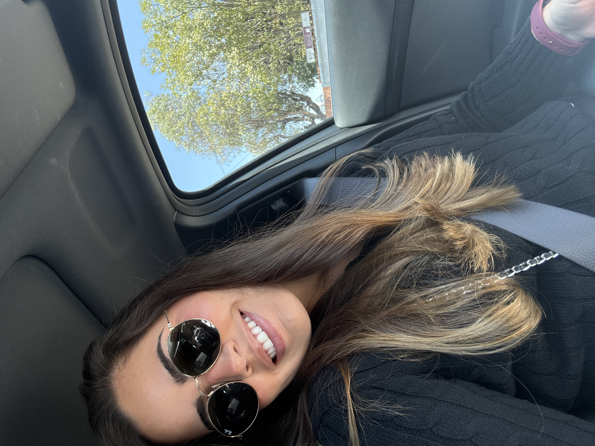 A female individual is featured in a close-up, chest-up selfie taken from inside a vehicle. She has long, dark brown hair with lighter, golden-brown highlights, styled over her shoulders. Her face displays a broad smile, revealing white teeth. She wears round, gold-rimmed sunglasses with dark lenses, reflecting a smartphone camera in the right lens. Her attire consists of a black, cable-knit sweater, and a gray seatbelt is fastened diagonally across her chest and left shoulder. A thin silver chain is visible near her left shoulder, and a dark pink or maroon wristband is present on her left wrist.
The vehicle's interior features a dark gray fabric headliner and a dark gray, textured plastic door panel. Through the passenger-side window, a clear blue sky is visible above numerous trees with vibrant green foliage. Below the trees, a light-colored wall or structure is present, with a partially obscured white rectangular sign showing dark text and a faint, triangular road sign in the background.
The background visible through the car window does not contain specific landmarks or legible street signage that would allow for the identification of a precise location within the city of Puebla, Mexico. The environment appears to be an outdoor setting, indicative of a sunny day in an urban or semi-urban area.