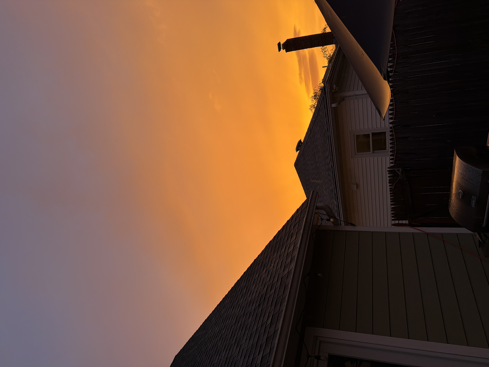 This is a nighttime photograph of a backyard in Riverhead, United States of America, during a vibrant sunset. The sky is a gradient of colors, transitioning from a soft purple at the top to a brilliant orange near the horizon. In the foreground, the dark roof of a house dominates the left side of the frame. To the right, a lighter-colored house with white siding and a window is visible, partially obscured by a dark wooden fence. A brick chimney extends upwards from the roof of the lighter house. In the lower right corner, the edge of a dark umbrella or canopy is visible. A barbecue grill is present in the lower portion of the image, its surface reflecting the warm light of the sunset. String lights are visible hanging from the gutter of the house on the left. The overall scene depicts a residential backyard at dusk with a dramatic sky.