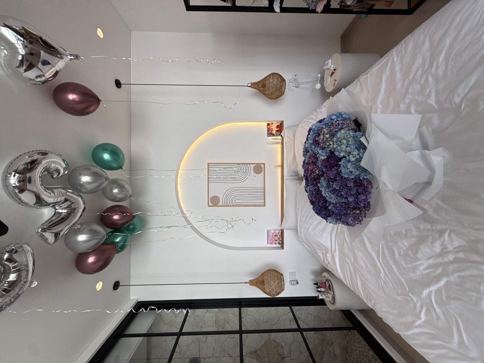 The image depicts the interior of a room decorated for a special occasion, potentially a birthday or anniversary. The location is likely in the Ubud District, Indonesia, given the architectural style and decorative elements.
The focus is on a bed covered with white linens, upon which a large bouquet of purple and blue hydrangeas is prominently displayed. To the left of the bed, balloons of various colors (silver, green, and purple) are floating, along with a large silver "25" balloon.
Decorative elements include woven pendant lights hanging from the ceiling, and a curved headboard with a modern geometric artwork. Small decorative items are placed on the bedside tables. Natural light enters the room through a large window, revealing what appears to be a stone exterior wall. No people are visible in the shot.