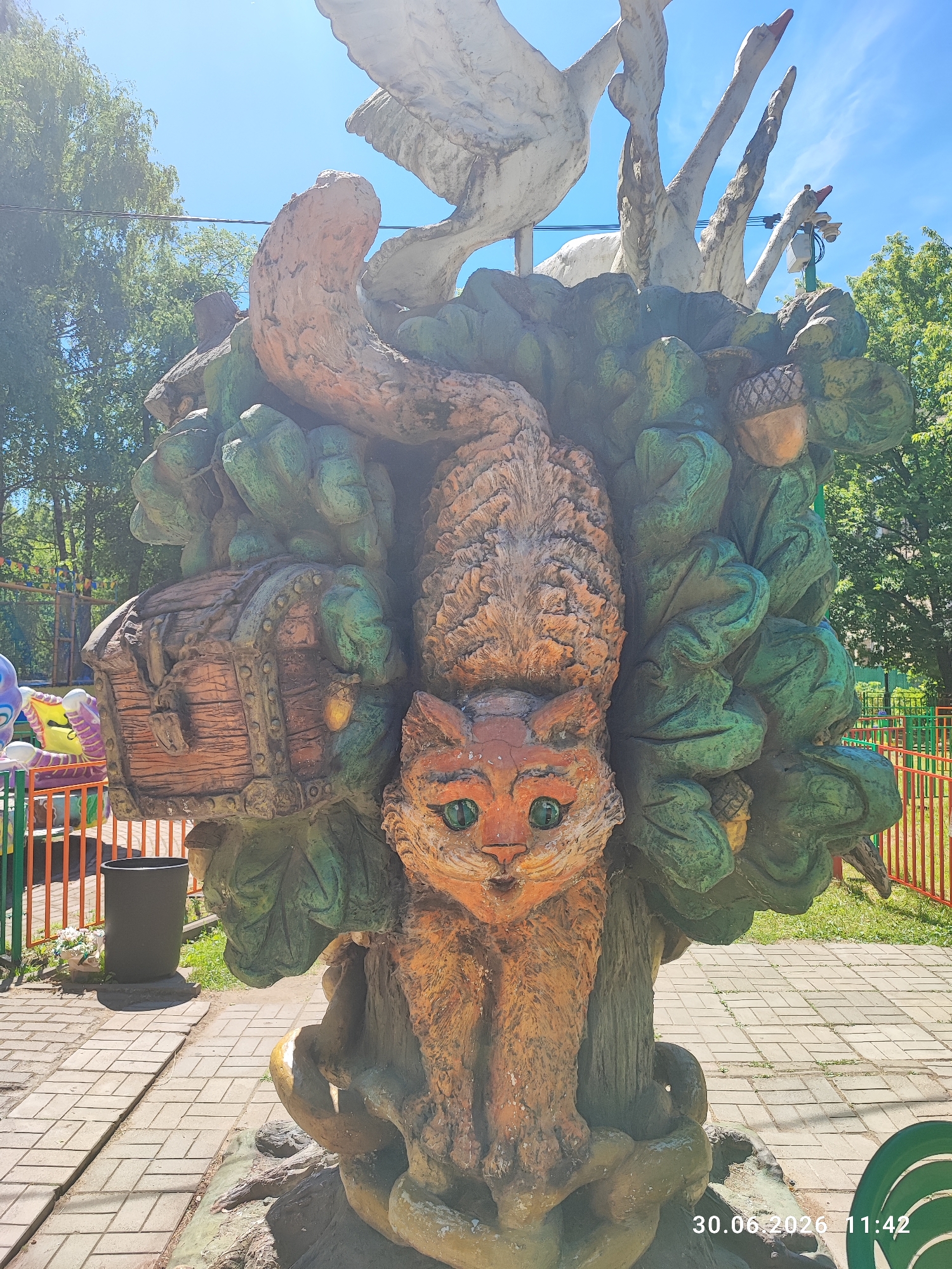 In Lytkarino, Russia, an outdoor sculpture depicts a whimsical scene centered around a large, orange cat-like creature with bright green eyes. The creature appears to be emerging from or intertwined with a cluster of large, green, leafy structures, resembling a tree or foliage. To the left of the cat's head, a wooden treasure chest with metal accents is attached to the sculpture. Above and behind the cat and foliage, stylized white bird-like figures with orange beaks are visible, suggesting a story or theme. The sculpture is situated on a paved area with a brick pattern, and in the background, there are fences, trees, and a bright blue sky. A trash can is visible to the left of the sculpture. The timestamp "30.06.2026 11:42" is visible in the bottom right corner of the image.
