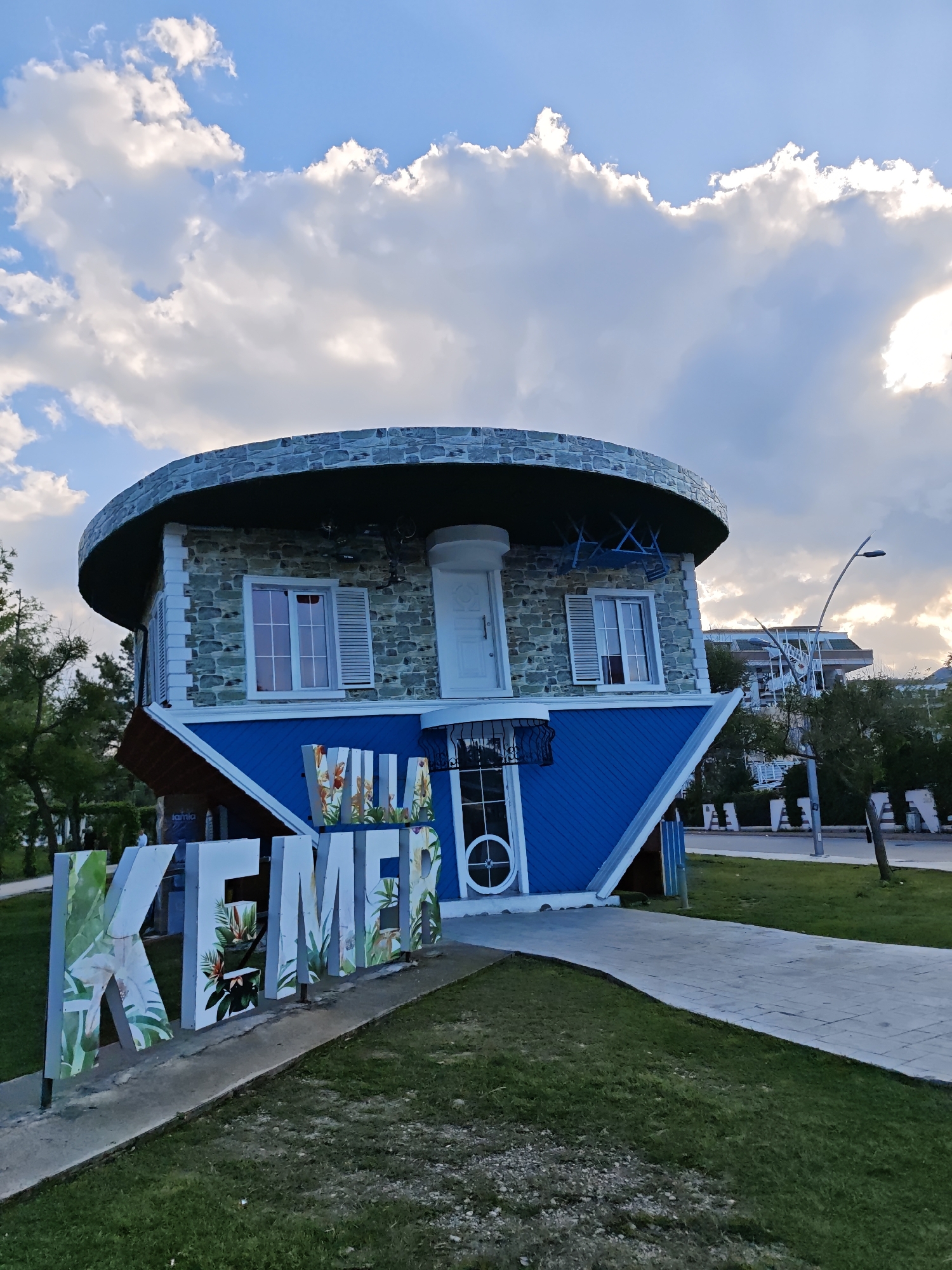 The image depicts an upside-down house tourist attraction in Kemer, Turkey. The house has a circular roof made of dark stone with a white door and two windows with shutters facing down. The exterior is a blue color. A sign spelling "KEMER" stands in front of the house, with each letter containing floral designs. A concrete walkway leads away from the house. The sky is cloudy with a hint of sunset light. In the background are trees and buildings, as well as street lighting.