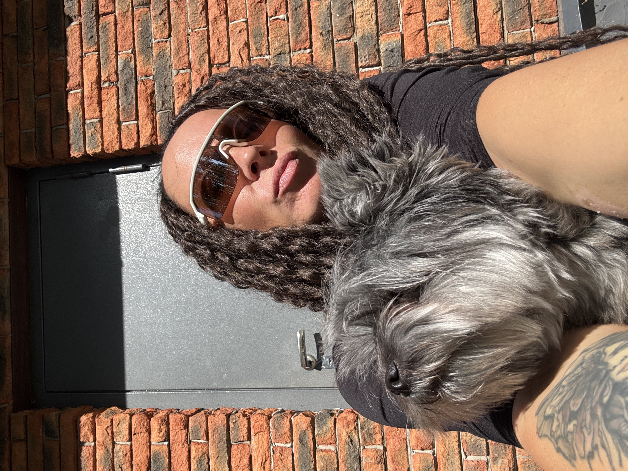 A person with dreadlocks and sunglasses is laying next to a gray, furry dog. The person has a tattoo on their arm. They are positioned in front of a red brick wall and a gray metal door with a latch. The location is not discernable as Chaikovsky, Russia.