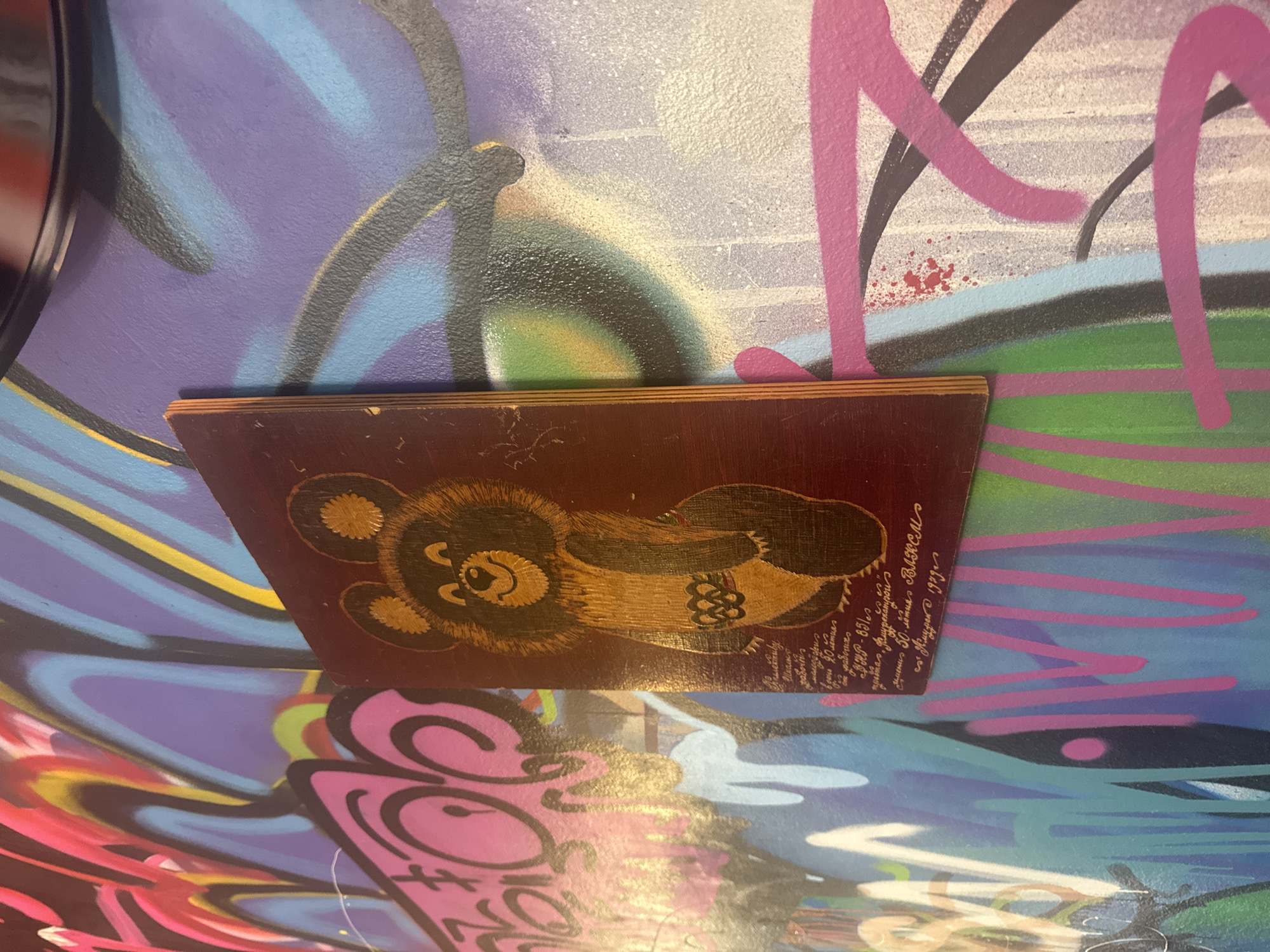 The image shows a close-up of a wooden plaque featuring a depiction of the Olympic bear, Misha, the mascot of the 1980 Summer Olympics. The plaque is affixed to a wall covered in vibrant graffiti. The bear is depicted in a traditional style, with a red background, and has the Olympic rings emblem on its chest. Below the bear, there is handwritten text in Cyrillic script. The graffiti on the wall is a mix of bright colors, including pink, purple, blue, green, and yellow, with stylized lettering and abstract shapes. The overall impression is one of urban art and cultural reference, with the plaque serving as a focal point against the backdrop of the graffiti. There are no people visible in the image, and no specific location within Komsomolsk-on-Amur, Russia, can be identified from the visual information alone.