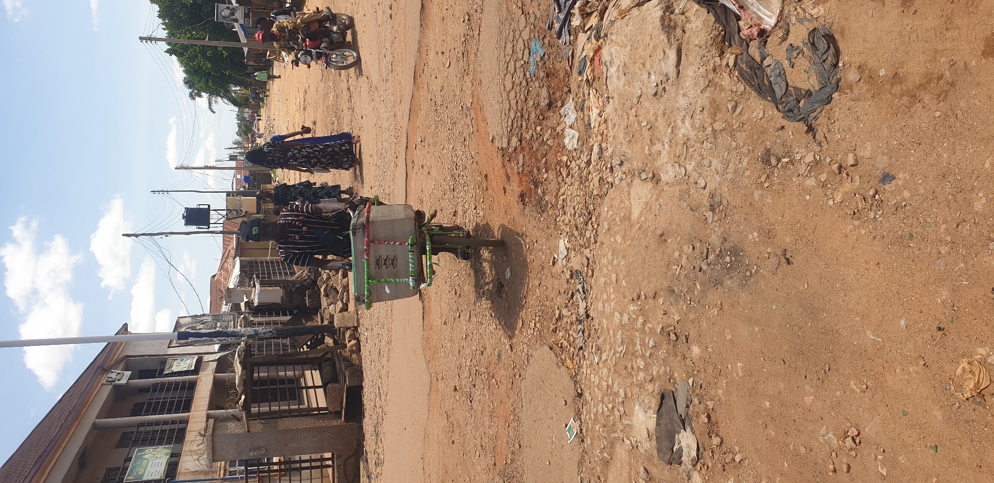 This image depicts a daytime scene on a dirt and gravel road in Sabon Tasha, Nigeria. The sky above is light blue with scattered white cumulus clouds, indicating sunny weather.
In the foreground, the reddish-brown ground is unpaved, uneven, and littered with rocks, small debris, and plastic waste. Visible potholes and eroded areas are present. The shadow of the primary subject extends across the ground.
The central subject is a male riding a bicycle towards the viewer. He wears a horizontally striped shirt (dark and light), dark trousers, a dark cap with a green emblem, and multiple beaded necklaces. The bicycle is equipped with a large, light-colored rectangular cooler or box mounted on the front. This box features green and red patterned ribbons or fabric around its edges, and text including "VALIANT MILK" with a cow logo is visible on its side.
Behind the cyclist, several individuals are present. To the right of the cyclist, a woman in a dark floral print long dress walks away from the camera, with another woman in a blue dress partially visible behind her. Further to the right, a red motorcycle with two occupants is approaching. The rider wears a red cap and light-colored traditional attire. In the background, additional pedestrians are visible walking along the street.
Buildings line both sides of the road. On the left, a multi-story building with a brown sloped roof and balconies features a green sign displaying "LADY'S CLASS BEAUTY SALON" and other text. Barbed wire is visible on a low wall in front of this building. On the right side of the street, single-story structures with corrugated metal roofs are visible. Multiple wooden utility poles with numerous overhead wires extend down both sides of the road. A black water tank on a metal stand is present in the mid-distance on the right. Sparse vegetation, including trees and bushes, can be seen beyond the buildings and further down the road.