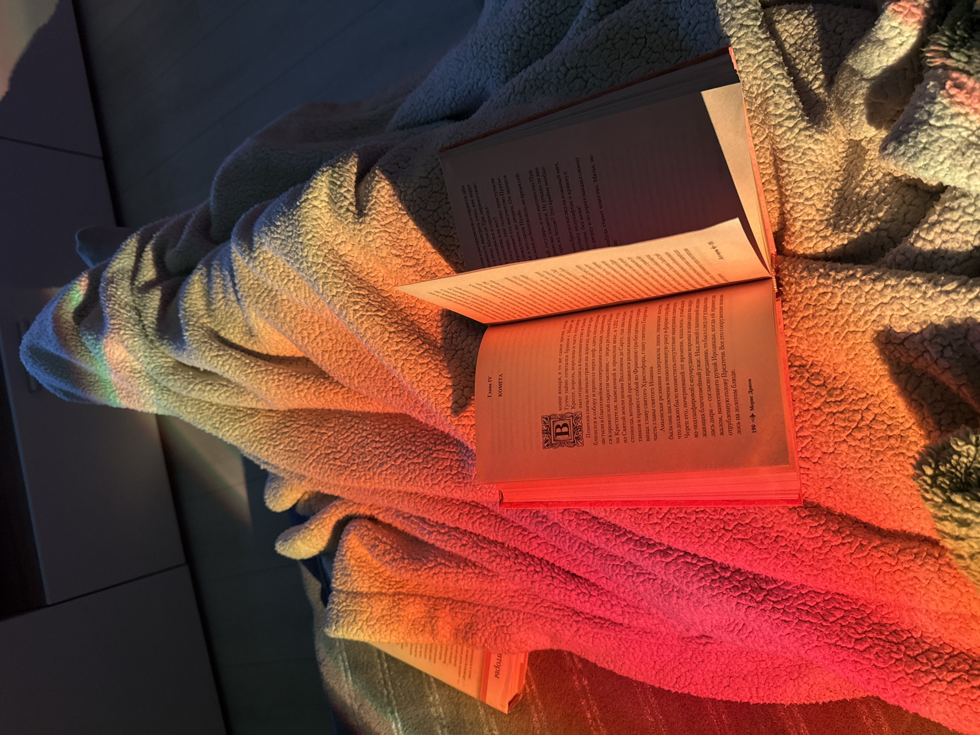 An open book rests on a soft, light-colored, textured blanket, illuminated by vibrant rainbow light. The scene suggests a tranquil moment of reading in a cozy indoor setting.
The most striking feature is the colorful illumination, which casts distinct bands of red, orange, yellow, green, and blue across the blanket's plush surface and the book's pages. This effect suggests sunlight passing through a prism or a similar refractive object, likely in the late afternoon or early morning given the warm tones. The blanket appears to be made of a fluffy, sherpa-like material, creating varied textures and shadows that are highlighted by the spectrum of light.
The book is open, displaying densely printed text in Russian. Visible details include "Глава IV KOMETA" (Chapter IV Comet) and "190 Морис Дрюон" (Maurice Druon), indicating the author and page number. Specific words like "Гуччо", "Матье де Три", "Пуатье", "Амьенская реликвия", "Христофора", "Георгия", "Иоанна", and "Иродиады" are discernible, hinting at historical or fictional narrative content. An ornate initial 'B' is also visible on one of the pages. The edges of the book's pages are particularly vibrant with red and orange hues from the ambient light.
The background shows a dark, indistinct indoor surface, possibly a wall or furniture, providing a subdued contrast to the brightly lit foreground. The overall atmosphere is one of serenity, comfort, and perhaps a touch of magic, created by the interplay of light, texture, and the implied activity of reading.