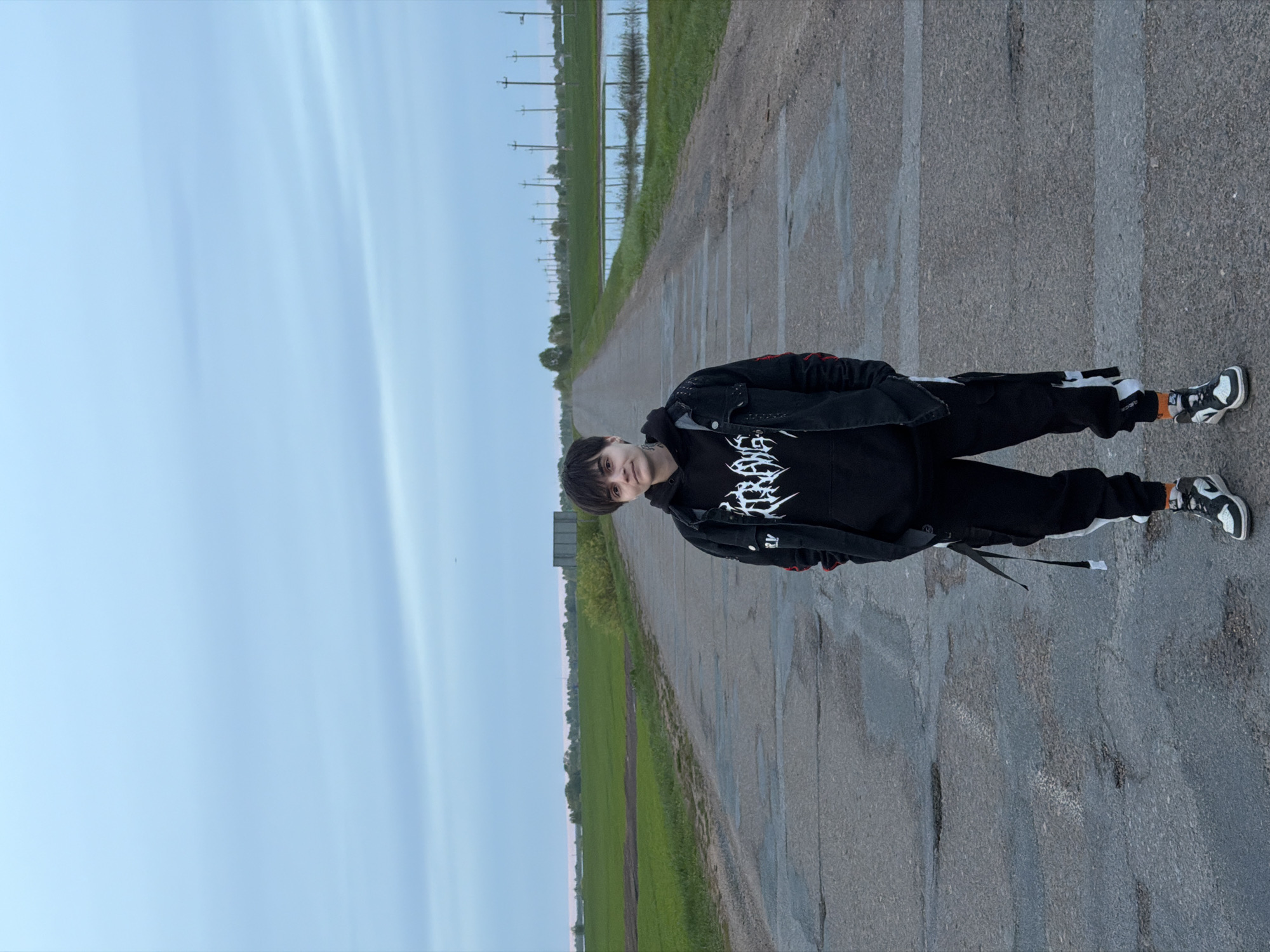 A person stands on a paved road in Azyakul', Russia, flanked by green fields and a distant line of utility poles. The person wears black clothing: a hoodie with a white graphic, a jacket, and pants. Their sneakers are black and white. The road stretches into the distance, reflecting the sky in some areas. The sky is pale blue.