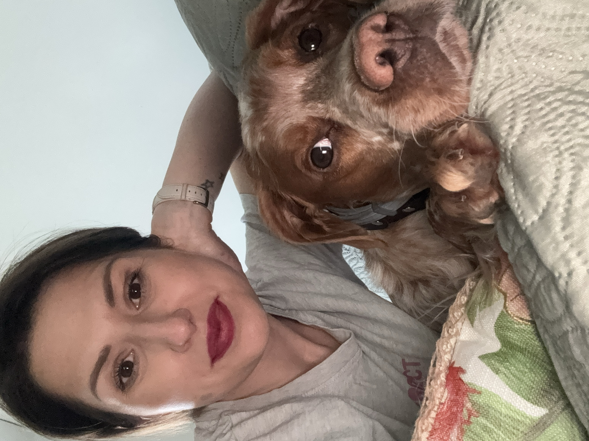 A selfie shows a woman and a brown dog lying on a patterned bedspread or blanket. The woman, on the left, is fair-skinned with dark hair and red lipstick. She wears a grey t-shirt and a watch on her left wrist. The brown dog, with large, expressive eyes, rests its head next to her. The background is a plain, light-colored wall. The location cannot be determined from the image.