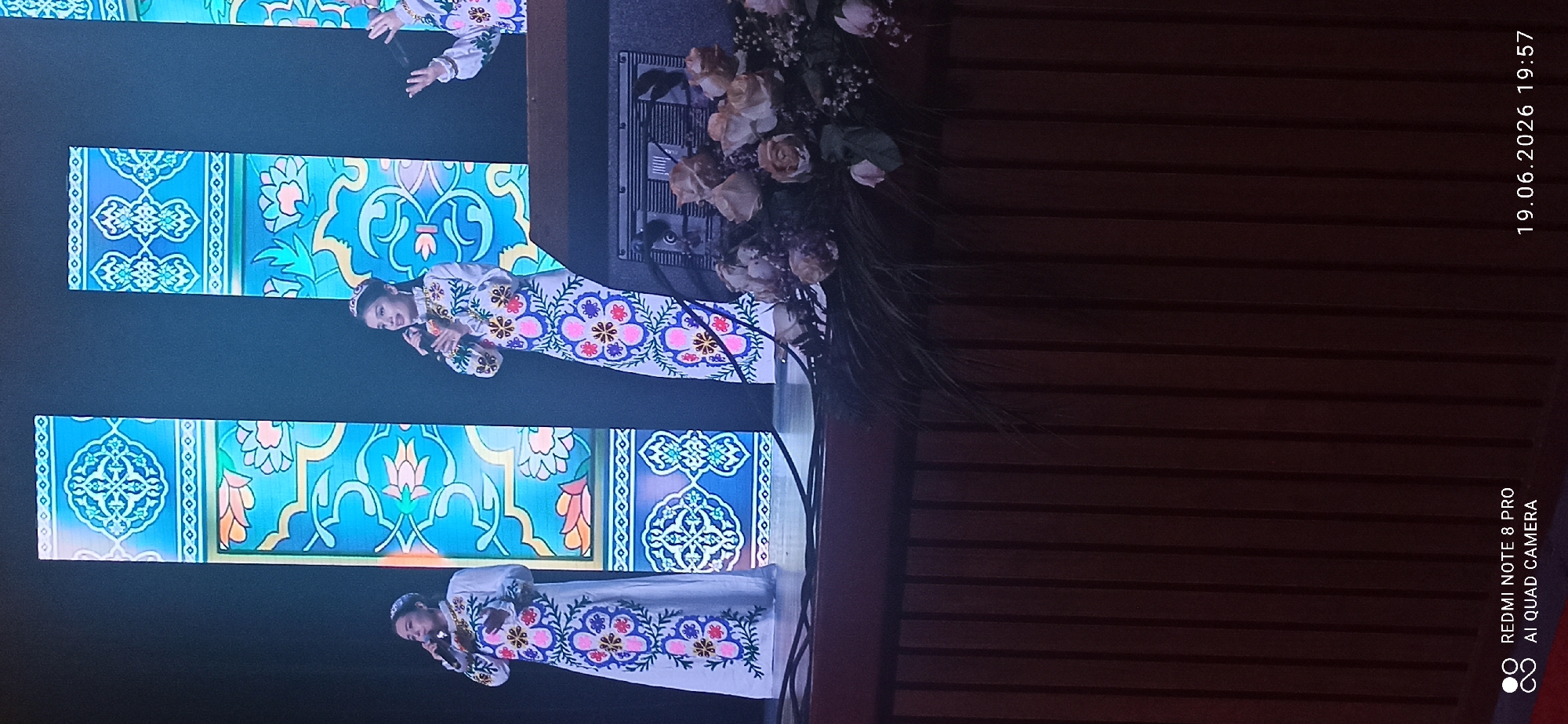 A stage is set with three young women in traditional, embroidered white dresses and headwear, holding microphones. They appear to be singing. The backdrop features large, illuminated panels with intricate blue and teal patterns. In the foreground, a floral arrangement sits on a dark wooden structure. The overall scene suggests a cultural performance or concert. The image was captured at an unknown time in an unknown city.
