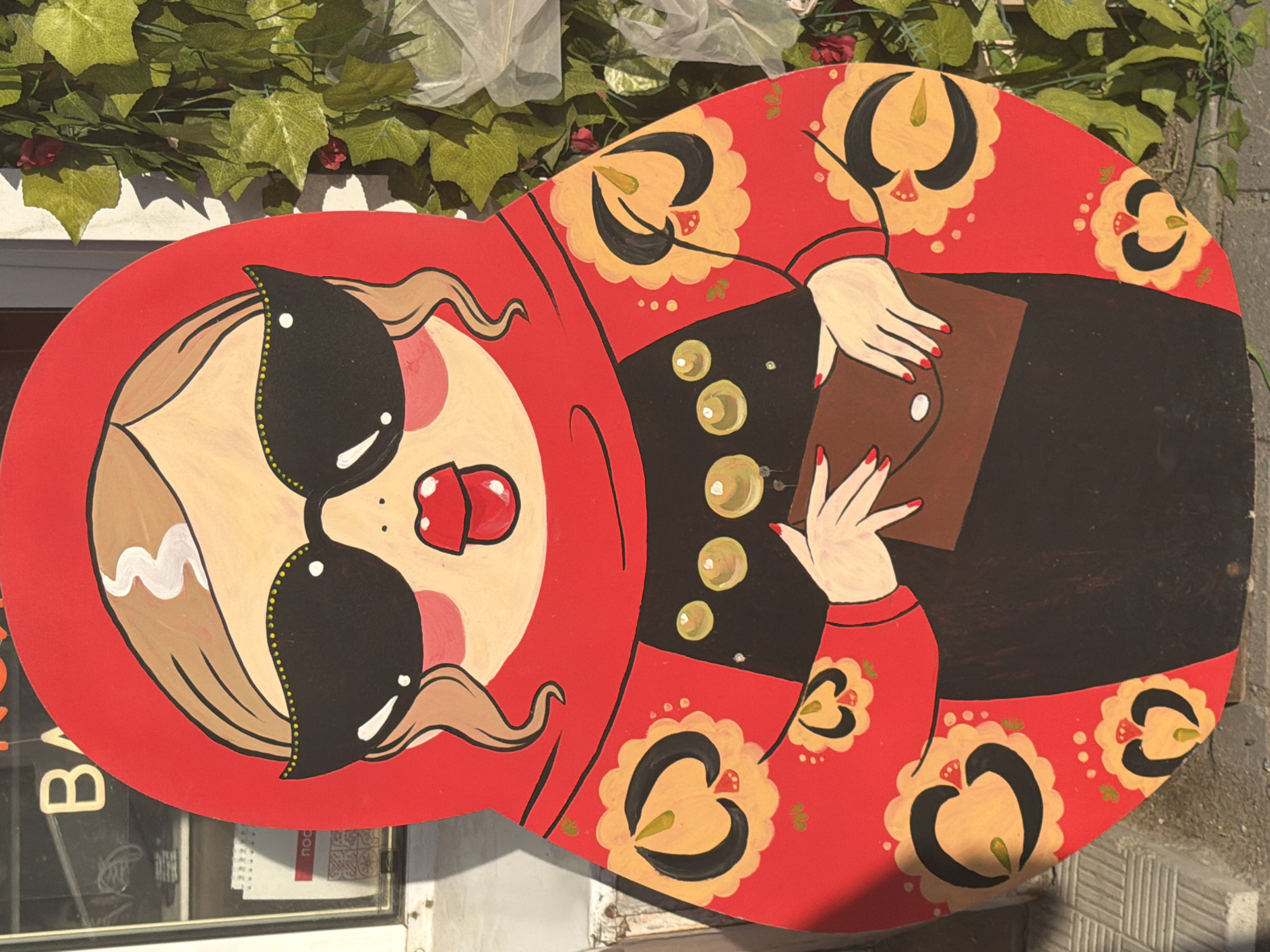 A vibrantly painted wooden matryoshka doll cutout, depicting a stylized woman wearing large black sunglasses and a red headscarf, is displayed outdoors. The doll has a red headscarf with a floral pattern on the red outer robe. She is holding a brown clutch bag. The cutout is positioned in front of a building with a window displaying some items and text that appears to read "BAG" and "NOC". Lush green ivy with red accents is growing on the right side of the image, behind the cutout. The ground beneath the cutout appears to be paved with gray textured material. This matryoshka doll cutout is located on the facade of a building in Vologda, Russia, specifically on Sovetsky Prospekt.