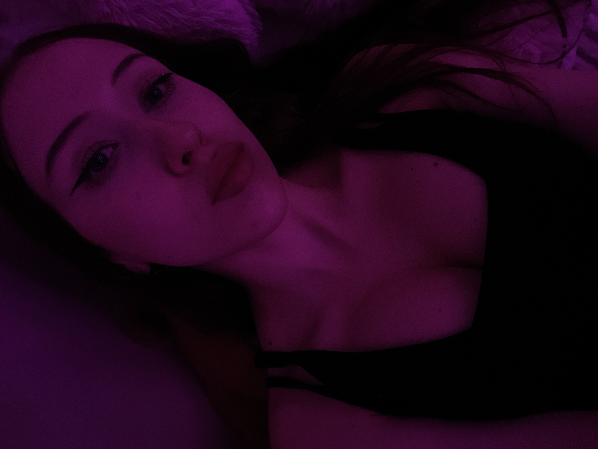 The image is a close-up, dimly lit selfie of a young woman. She is wearing a black top. The lighting is predominantly a vibrant purple hue, casting a strong glow on her face and upper body. Her makeup includes winged eyeliner. The background is dark and indistinct, with a suggestion of a textured surface, possibly fur or fabric, to her right. There is no visible indication of the city of Votkinsk, Russia, or any specific environmental context within the image.
