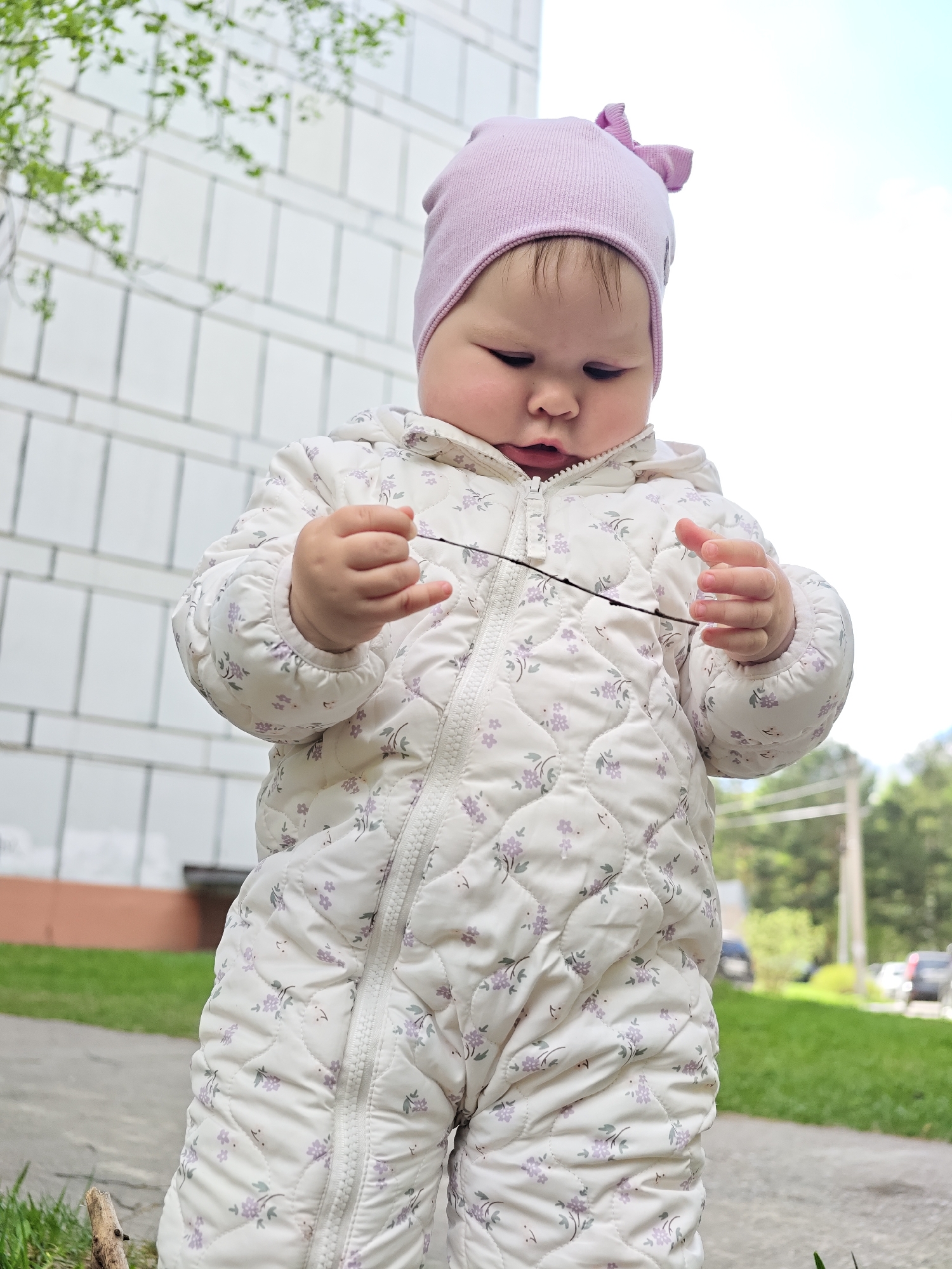 A baby stands outdoors on a concrete surface next to a patch of green grass. The baby is wearing a light purple hat with a bow, and a white quilted snowsuit with a small floral pattern. The baby is holding a thin, dark twig between its hands, looking down at it with focused attention. Behind the baby is a white brick building, a blurred view of trees, and parked cars. The location is Chaikovsky, Russia.