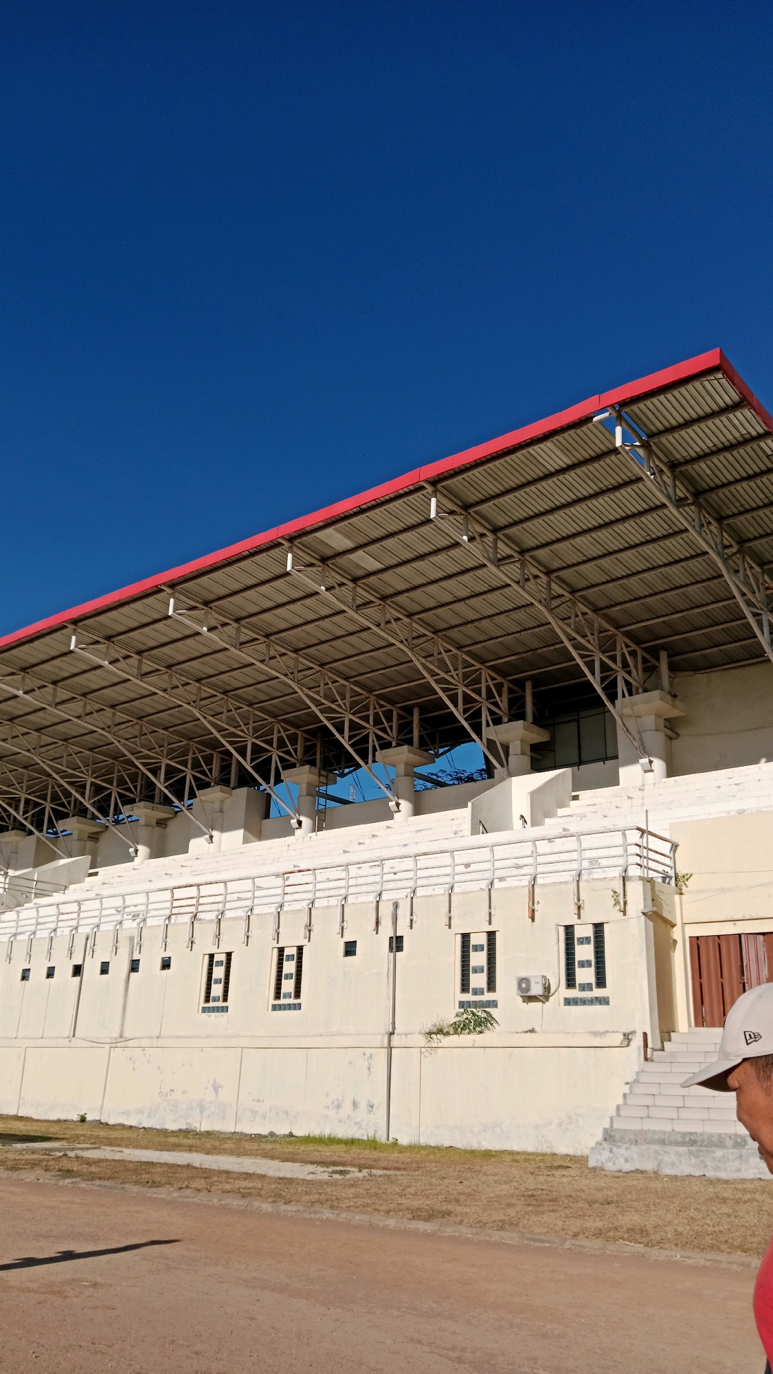 The image depicts a clear, bright blue sky above a stadium grandstand. The grandstand has a metal roof with a red trim, supported by a complex network of metal beams and columns. Below the roof, the grandstand itself is made of concrete and stone, with rows of white seating visible. The front of the grandstand features a series of windows and what appear to be ventilation units. In the foreground, a dirt track runs horizontally across the image. To the right, a portion of a person wearing a white baseball cap and a red shirt is visible, looking towards the stadium. The scene appears to be outdoors, in a sunny environment. The image captures a section of the stadium located in Mangkujayan, Indonesia.
