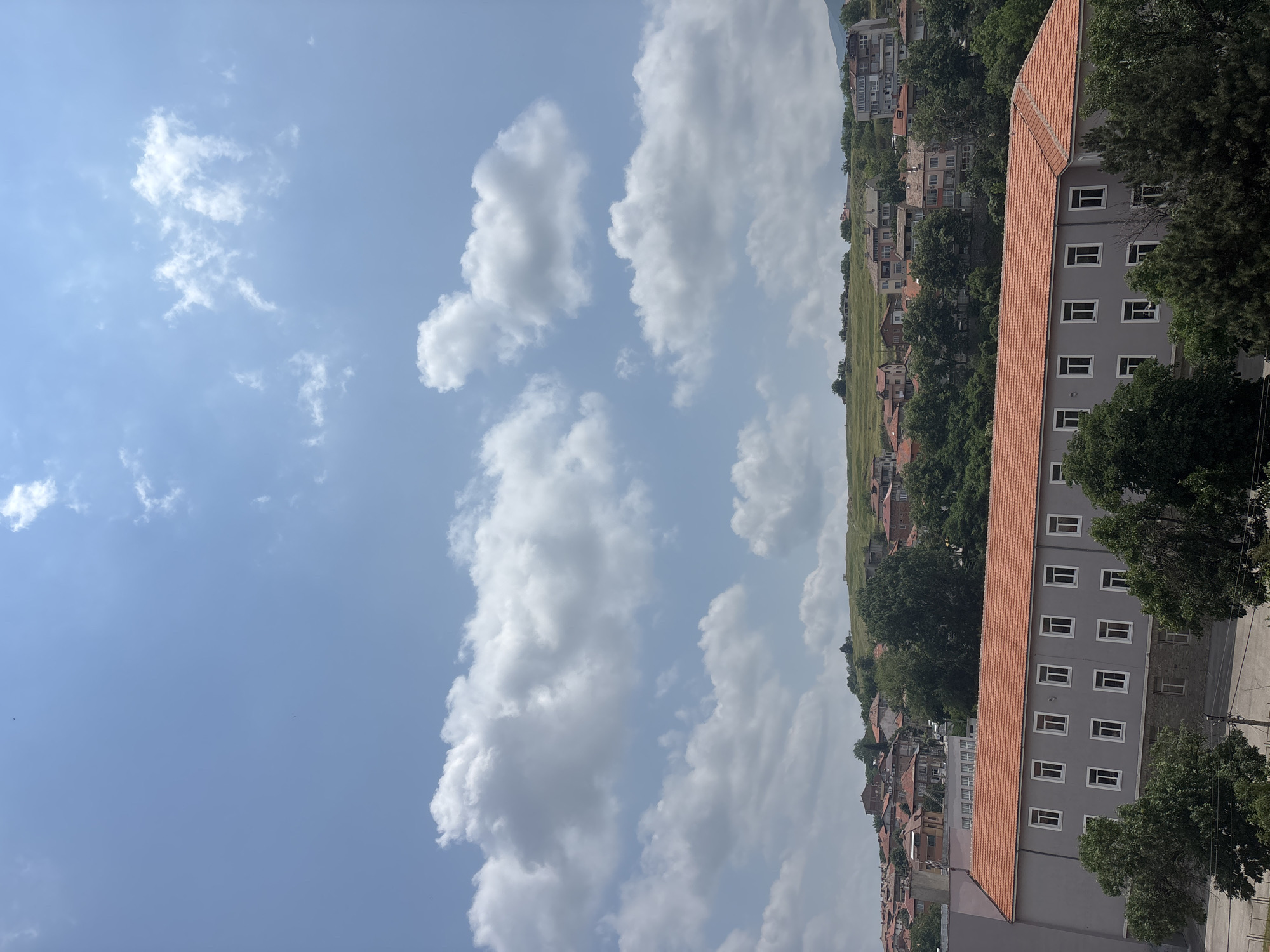 The image presents a panoramic view of a residential area in Asenovgrad, Bulgaria, under a clear, bright sky. The perspective suggests the photo was taken from an elevated position, looking down across a valley or hillside dotted with buildings.
The upper portion of the image is dominated by a vibrant blue sky, sparsely populated with fluffy white cumulus clouds, indicating fair weather. The sunlight is strong, suggesting it is daytime, likely late morning or early afternoon.
The mid-ground and foreground feature a dense arrangement of residential buildings. On the right side, a prominent, long, grey-facaded building with numerous rectangular white-framed windows and a distinct reddish-orange tiled roof stretches horizontally across the frame. Surrounding this building and extending into the middle distance are various other houses and structures, many also featuring traditional red-tiled roofs. The architecture varies, with some buildings appearing more modern and others exhibiting older styles common to the region.
Greenery is interspersed throughout the urban landscape, with numerous trees providing shade and breaking up the uniformity of the rooftops. A large, verdant, grassy hill or field is visible in the mid-ground, rising behind the cluster of buildings. In the far distance, faint outlines of mountains or higher hills can be discerned, partially obscured by a light haze or distance. There are no people or specific activities visible, making it a tranquil scene of an everyday Bulgarian townscape. No visible text is present in the image.