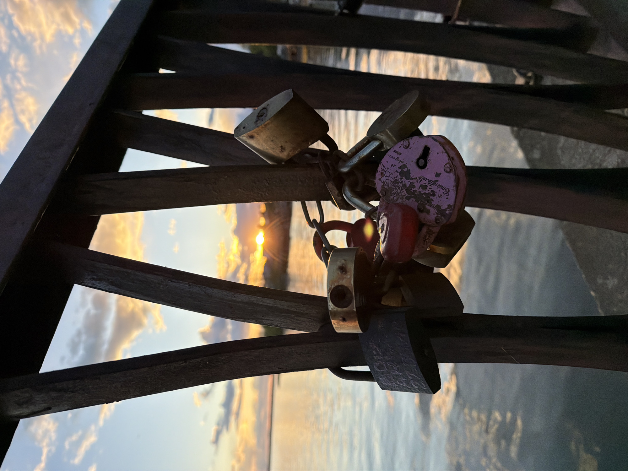 The image depicts a cluster of padlocks attached to a dark wooden structure, likely a bridge or railing. The locks are of various sizes, colors, and materials, with a notable pink padlock and a chain linking several together. The background shows a body of water reflecting the light of a setting or rising sun, illuminating the sky with warm hues and scattered clouds.
The location is presumed to be in Votkinsk, Russia, although this is not evident within the image itself.