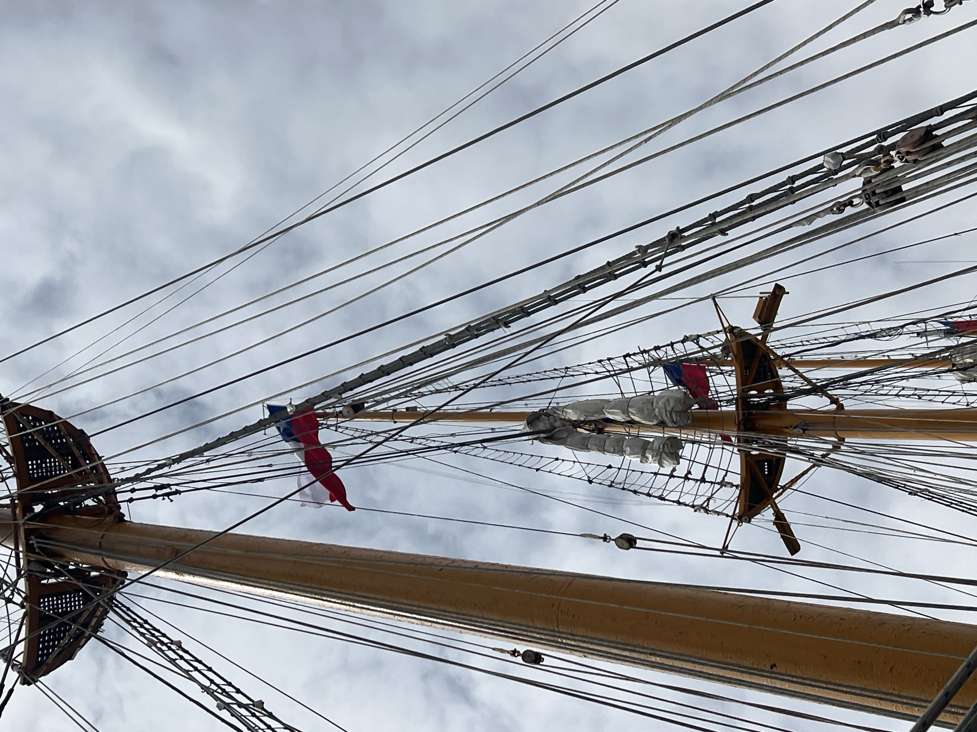 The image shows the masts, rigging, and yardarms of a tall ship against a cloudy sky, likely taken in Arica, Chile. Two flags are visible on the masts. The masts are painted yellow, and the rigging is a complex network of ropes and lines. There are wooden structures, including a crow's nest, visible on the masts. The background is a cloudy sky. The scene suggests the tall ship is docked or sailing near Arica, Chile, given the context provided.