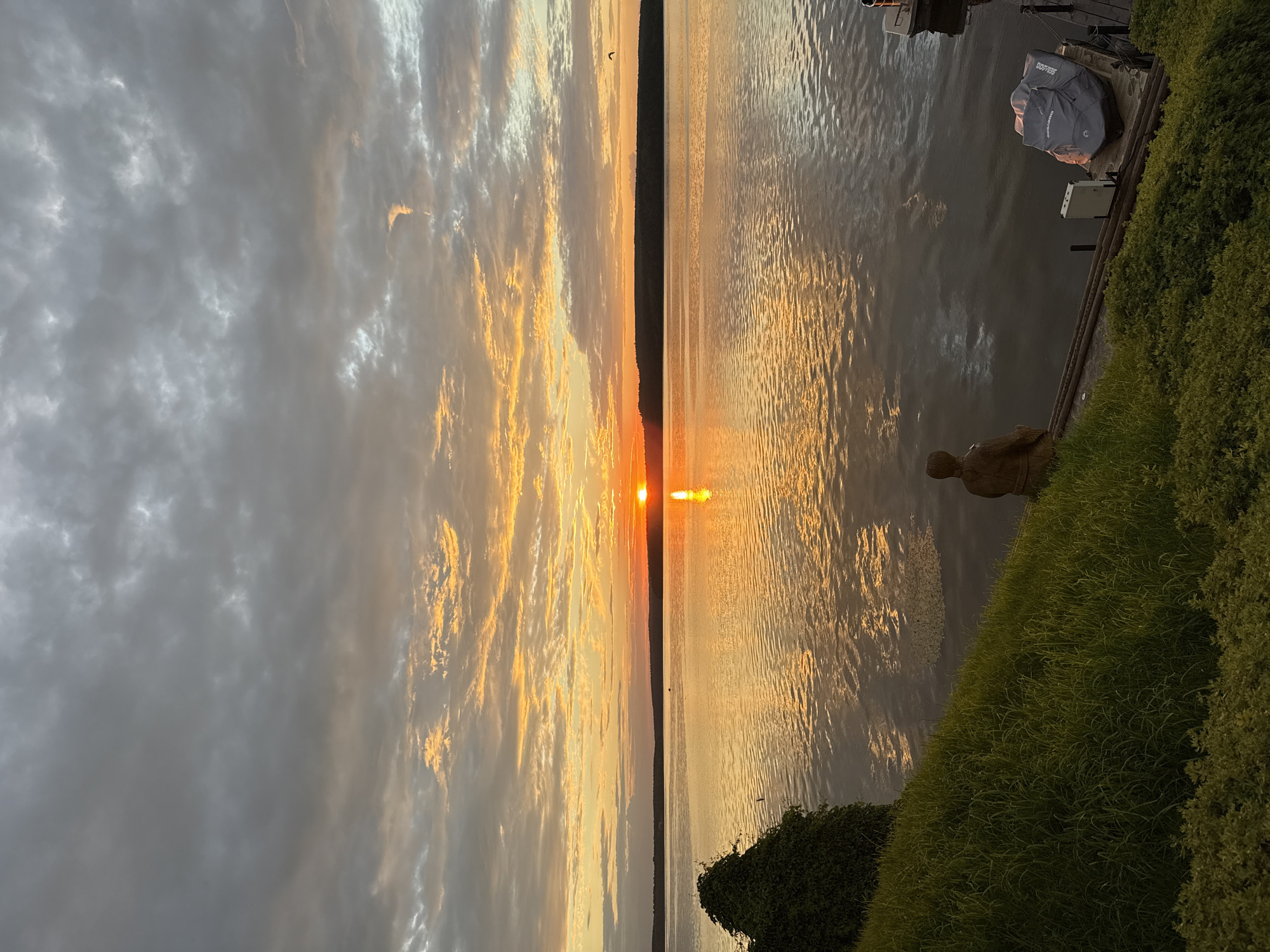 The image depicts a serene sunset over a body of water, presumably the Kama River, as seen from Votkinsk, Russia. The sky is filled with dramatic clouds, tinged with shades of orange and yellow from the setting sun. The sun itself is a bright orange orb, casting a shimmering reflection on the calm water. In the foreground, a grassy bank slopes down to the water's edge. A small, dark statue of a person is seated on the bank, gazing out at the sunset. A jet ski is parked on a small dock to the right of the statue. The overall impression is one of peaceful contemplation and natural beauty.