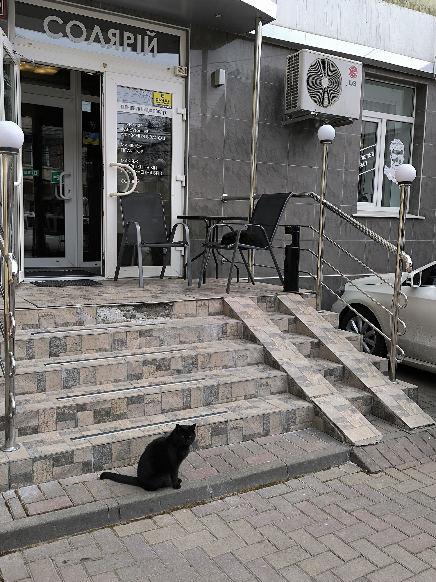Outside a business labeled "СОЛЯРІЙ" (solarium) in Vinnytsia, Ukraine, a black cat sits on the curb in front of the tiled steps leading to the entrance. The steps have a ramp on either side. Two chairs and a table are on the landing outside the door. Above, there's an LG air conditioning unit and a window with a barber-style illustration. A silver car is parked to the right. The establishment offers over 70 types of services including haircutting, hair coloring, hair treatment, manicure, pedicure, makeup, eyelash extensions, eyebrow shaping, and tanning.