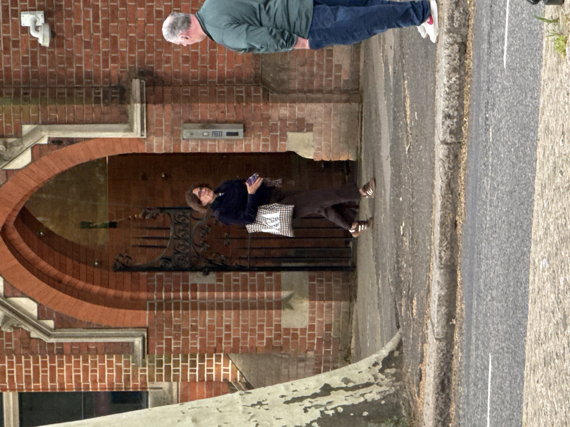 The image depicts a street scene. A woman with dark hair and glasses stands on the sidewalk, holding a mobile phone. She wears a dark jacket, brown pants, and sandals, with a black and white checkered bag. Behind her is a brick building with an arched entryway, secured by an ornate wrought iron gate. Above, a man with gray hair is partially visible, wearing a green sweater and jeans. The sidewalk is concrete, and the street has a white line marking. A surveillance camera is mounted on the building. Location is likely in London, United Kingdom given the architectural style.