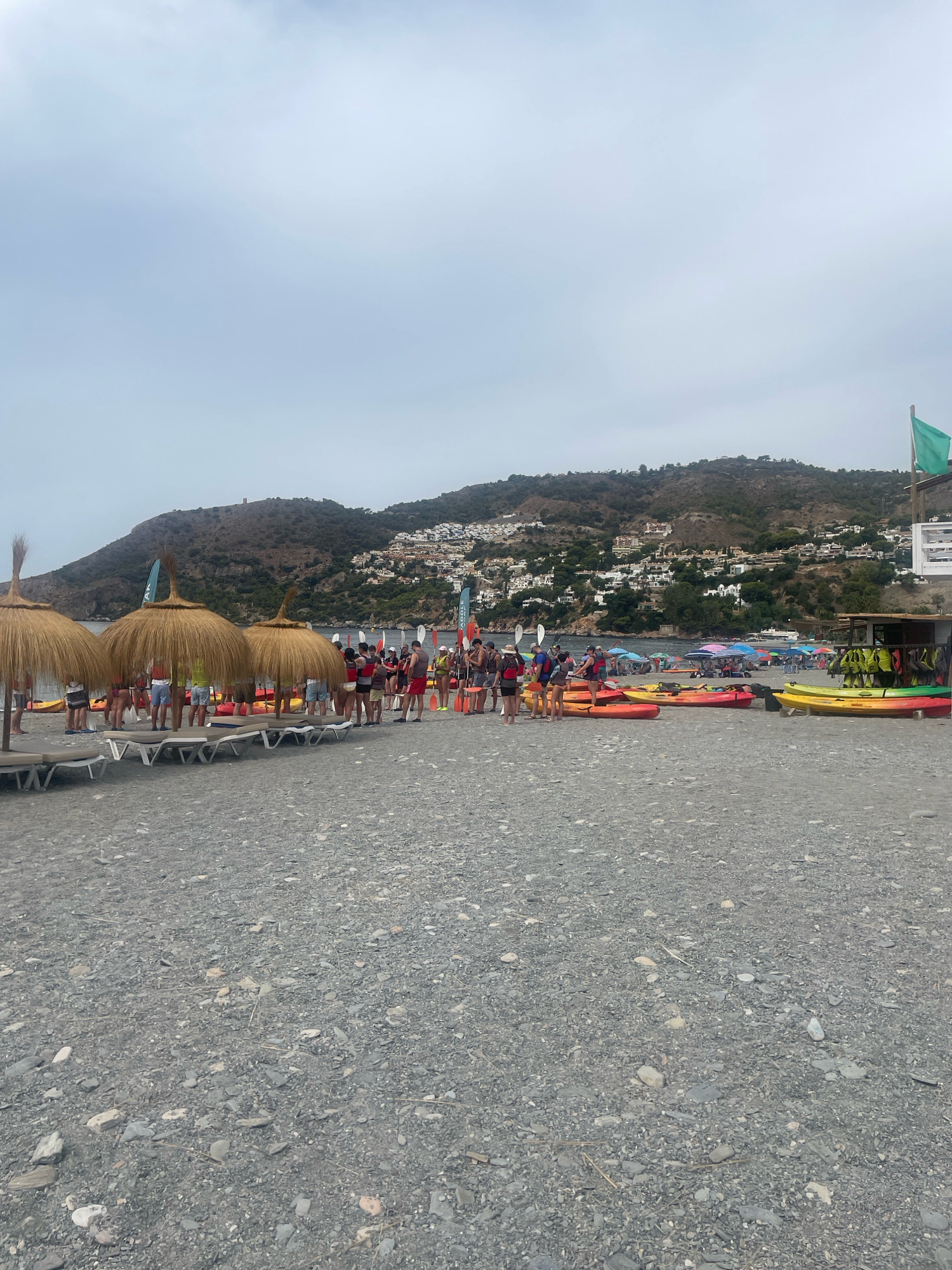The image shows a crowded beach in La Herradura, Spain, with many people gathered around kayaks and inflatable boats. A group of individuals, likely tourists, are wearing life jackets and appear to be preparing for a water activity. Several thatched umbrellas and beach chairs are set up on the pebbly shore. In the background, a hillside town with white buildings is visible, cascading down towards the sea. The sky is overcast, and the overall atmosphere suggests a busy recreational day at the beach.