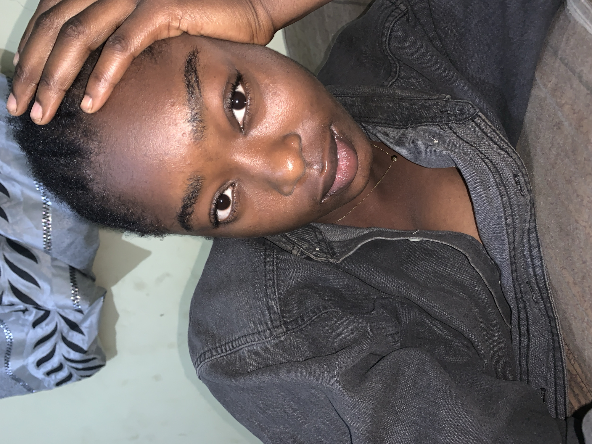 A person with dark skin and short, dark hair is posing for a photo indoors. The person is wearing a dark gray denim jacket and a thin gold necklace. Their right hand is touching their forehead. The background includes a light green or beige wall and a piece of silver and black patterned fabric or clothing. The location in Rafin Par, Nigeria, is not discernible from the image.
