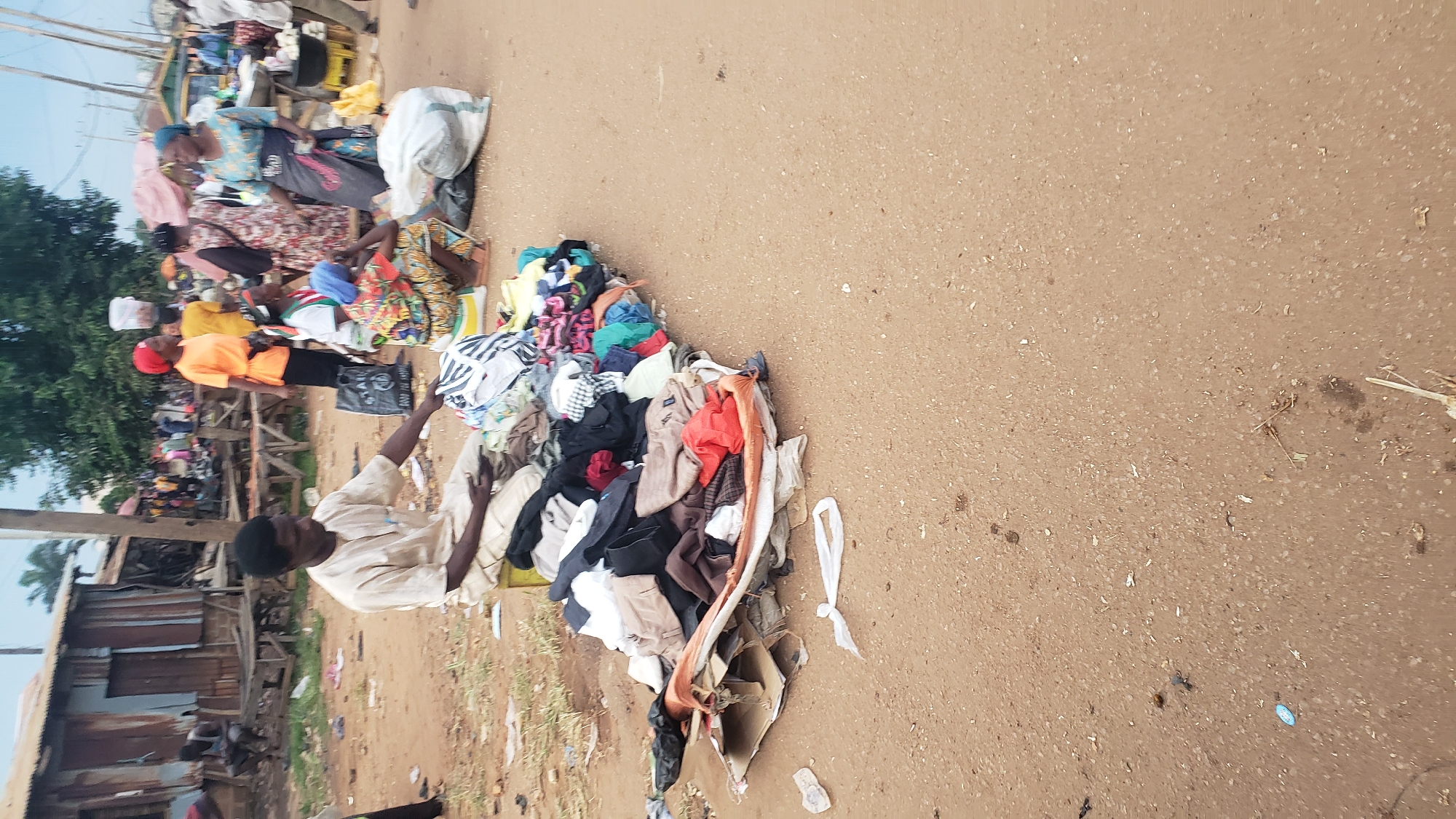 In the city of Kakuri Gwari, Nigeria, a scene unfolds at a roadside market. In the foreground, a man dressed in a beige tunic and trousers sits on a yellow crate, his attention focused on a pile of clothing spread out on the ground before him. The clothing, a jumble of various colors and patterns, is arranged on top of a makeshift display of cardboard and fabric. To his right, a group of individuals are gathered, some standing and others seated. One woman wears a bright orange shirt and a red headscarf, while another is adorned in a blue and white patterned dress. A woman in a colorful patterned dress is seated, wearing a blue head covering. Behind them, a more extensive market is visible, with stalls and people milling about. The ground is a mixture of sand and gravel, indicative of an outdoor urban setting.