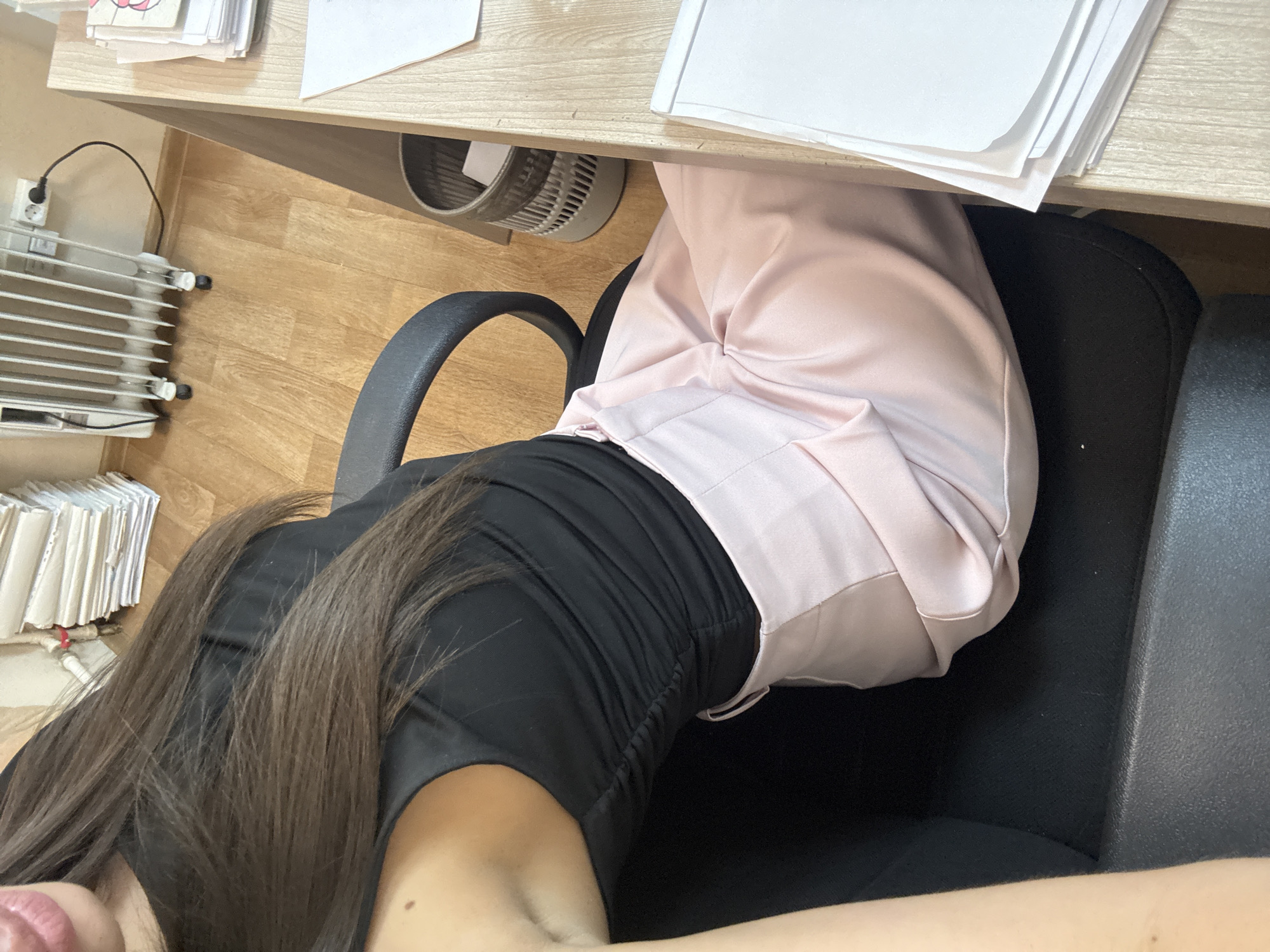 The media file shows a person sitting at a desk. The subject is wearing a black top and pink pants. Their long brown hair is visible, as are their shoulders and neck. The scene is an indoor office or study setting. On the desk, there are stacks of papers, some of which are in folders. A small, cylindrical fan is visible under the desk, and a white electric heater is plugged into the wall on the wooden floor. The lighting suggests it could be daytime, but the specific time of day or weather conditions cannot be determined. No text is clearly visible in the image.