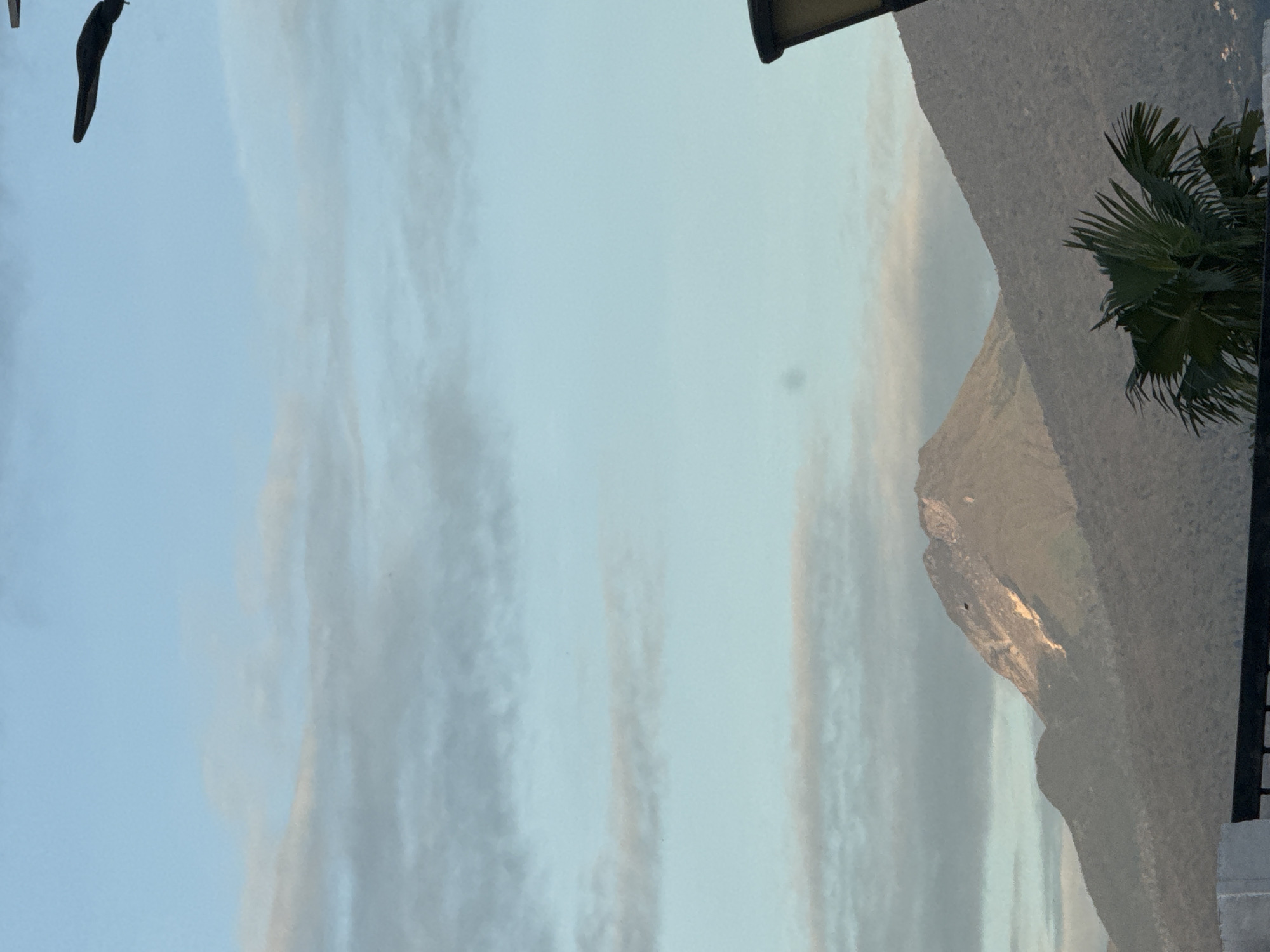 The image shows a view of a mountain range from what appears to be a residential or commercial building. A palm tree frond is visible in the bottom right corner, suggesting a tropical climate. The sky is partly cloudy with soft, muted colors. The mountain's rocky terrain is illuminated by the sun, casting shadows on its surface. The location is in the city of Davao City, Philippines.