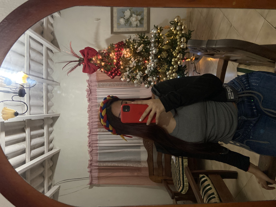 A mirror selfie shows a woman holding a red phone, partially obscuring her face. She wears a braided, colorful headband. Reflected in the mirror is a Christmas tree decorated with lights and ornaments. Also visible are light pink curtains, a framed picture of white flowers, and a chandelier. The woman wears a gray shirt and a black jacket, along with embellished jeans. The reflection shows a wooden chair with striped cushions.