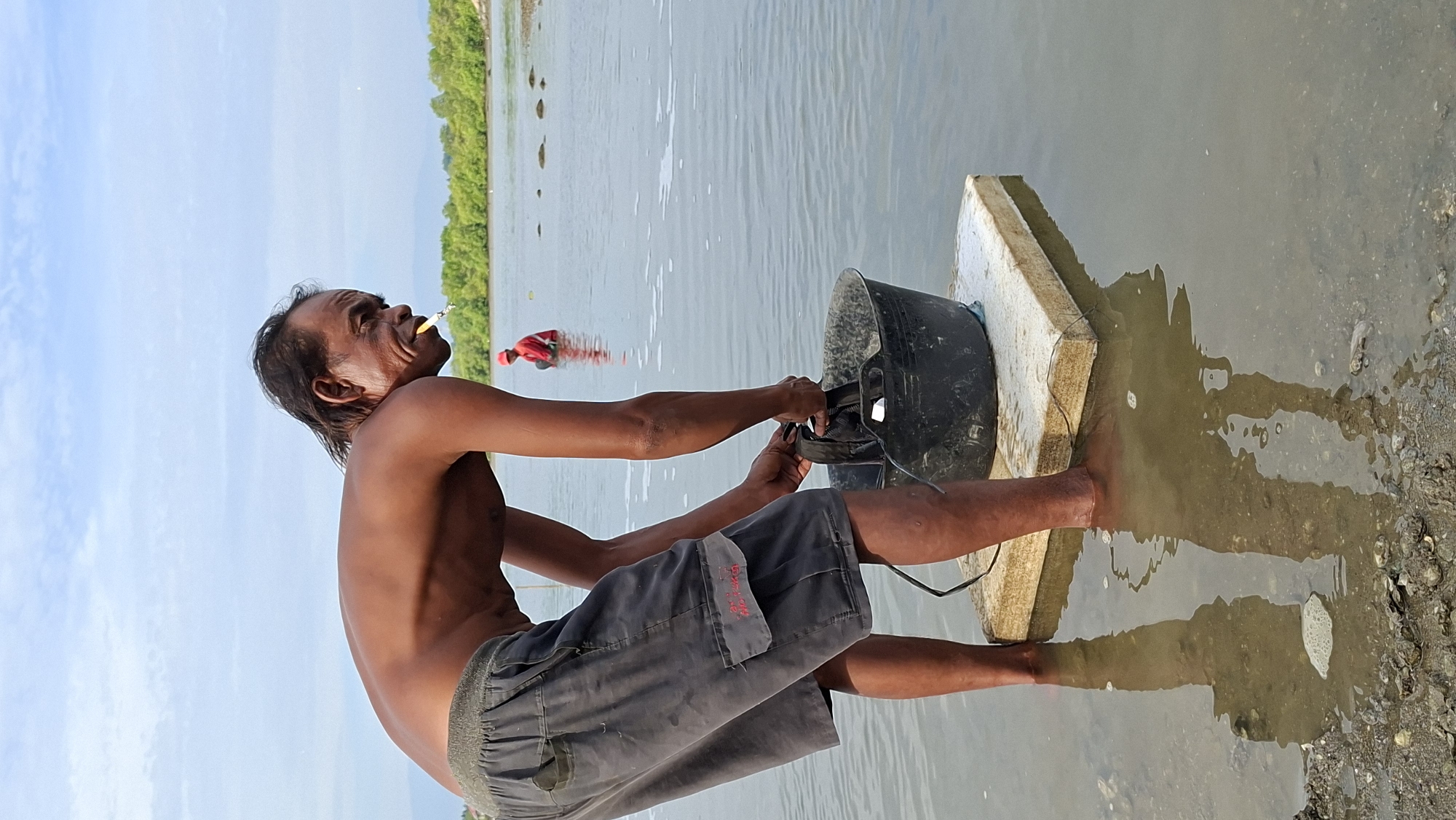 An adult male with dark skin and shoulder-length dark hair is standing in shallow water, bending over to handle a black bucket. He is wearing dark gray shorts with a red embroidered pocket detail and appears to be smoking a cigarette, which is visible in his mouth. He is standing on a rectangular white foam object. In the background, another person in a red shirt is partially submerged in the water. The environment is a body of water with a shoreline visible in the distance, characterized by green foliage and a light blue sky with scattered clouds. The water is murky and appears to be a coastal or estuarine area. The image was captured outdoors in Lhokseumawe, Indonesia.