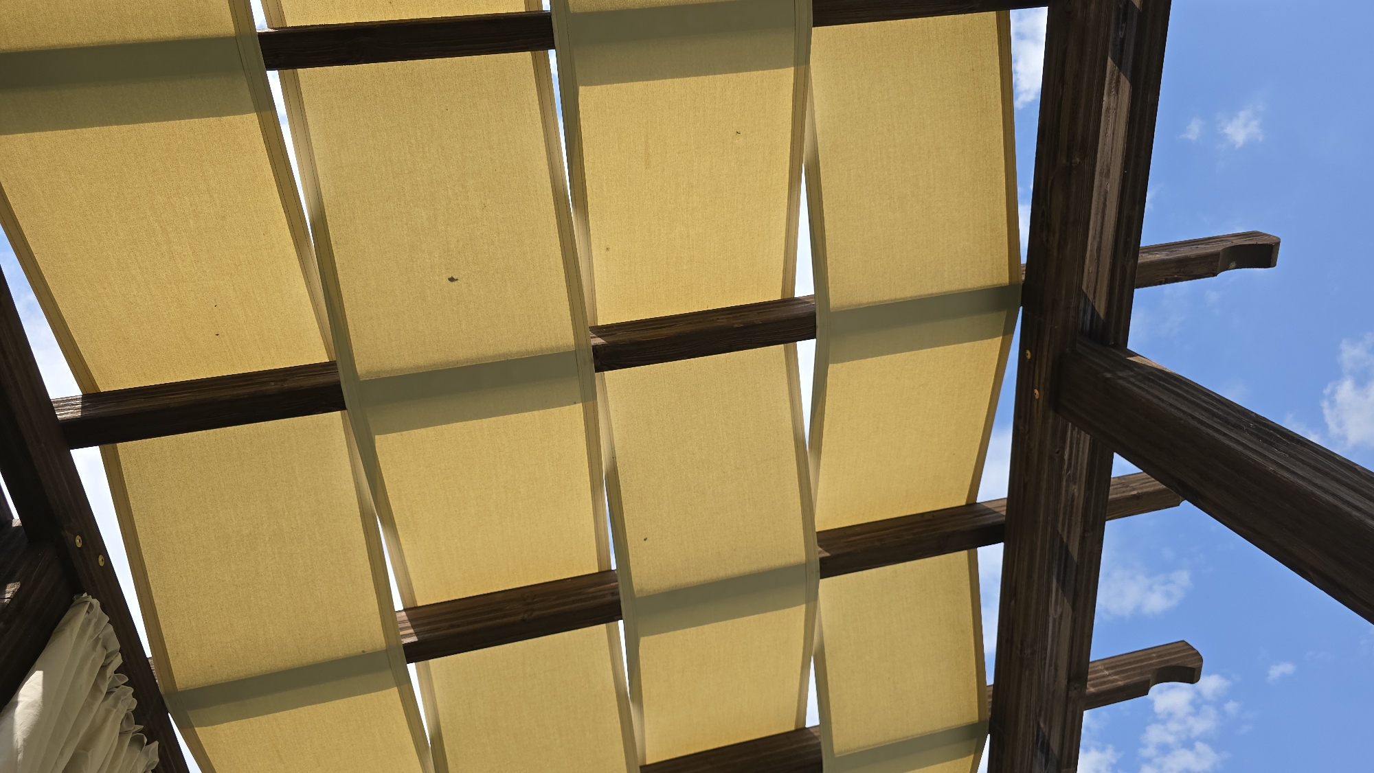 The image shows a close-up, upward view of a wooden pergola with yellow fabric shades. The pergola's dark brown, weathered wooden beams crisscross, supporting rectangular panels of yellow fabric. The fabric is stretched taut, creating a patterned effect with alternating sections of light and shadow, suggesting sunlight filtering through. Gaps between the fabric panels reveal a bright blue sky with scattered white clouds. A small, dark speck, possibly an insect, is visible on one of the fabric panels. In the upper left corner, a white pleated fabric is also visible, possibly part of a retractable awning. The scene appears to be outdoors on a sunny day, likely in a garden or outdoor seating area. The image is from Krasnovo, Bulgaria.