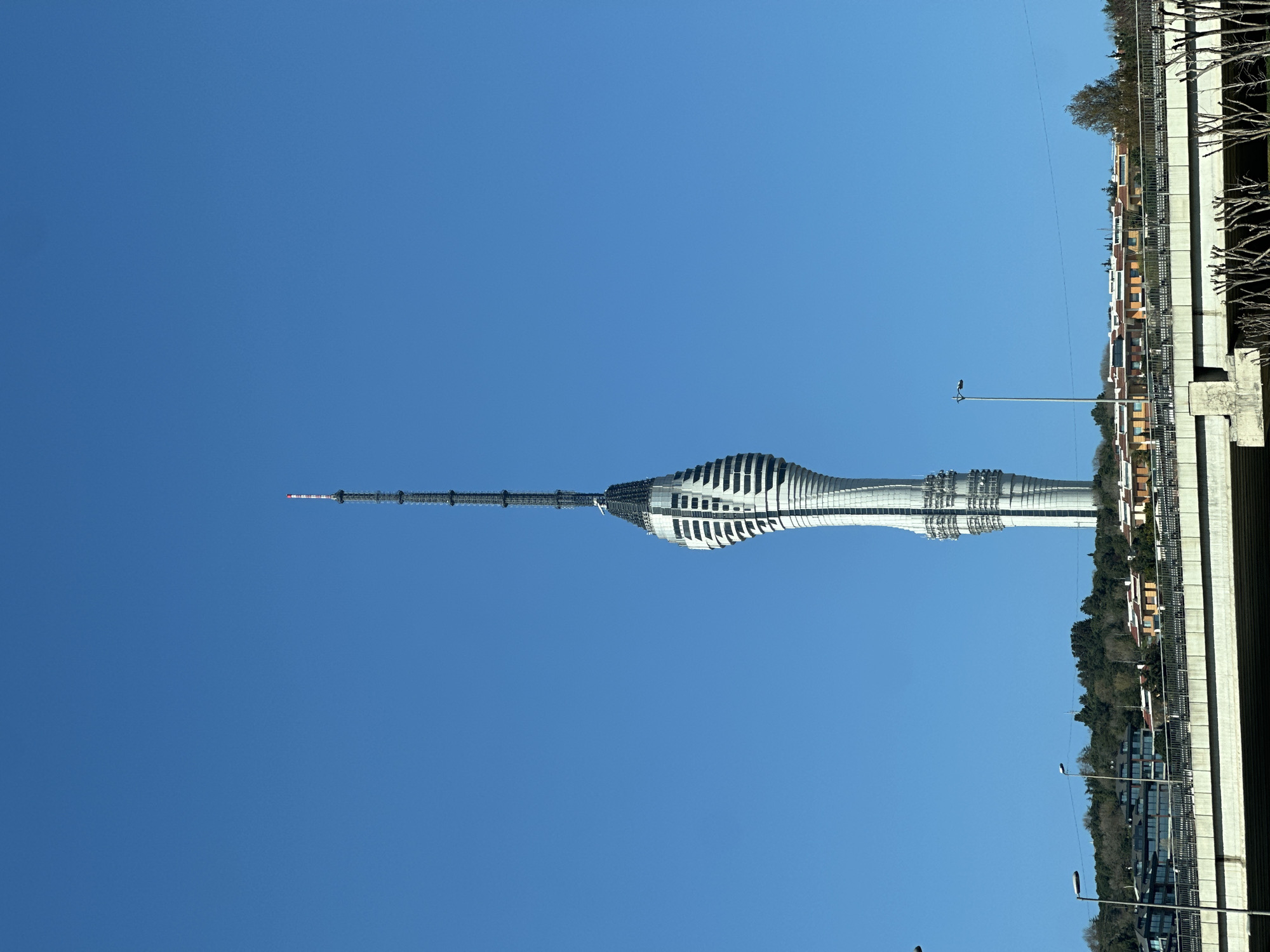 The image shows the Çamlıca Tower in Üsküdar, Turkey, against a clear blue sky. The tower is a modern structure with a distinctive, layered design. The base of the tower is wider, narrowing as it ascends before expanding again near the top. The tower is silver and white in color. A long, needle-like antenna extends from the top of the tower. To the right of the tower are buildings and trees, suggesting the presence of a city or urban area. The perspective is from below, looking upwards at the tower. The image is rotated 90 degrees counterclockwise.