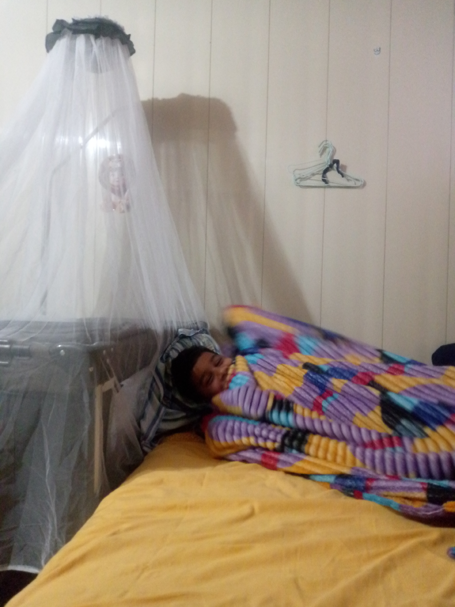 A child lies on a yellow bed with a colorful blanket, possibly in a bedroom in Altagracia, Venezuela. A mosquito net hangs over the bed's edge. Empty clothes hangers are on the wall.