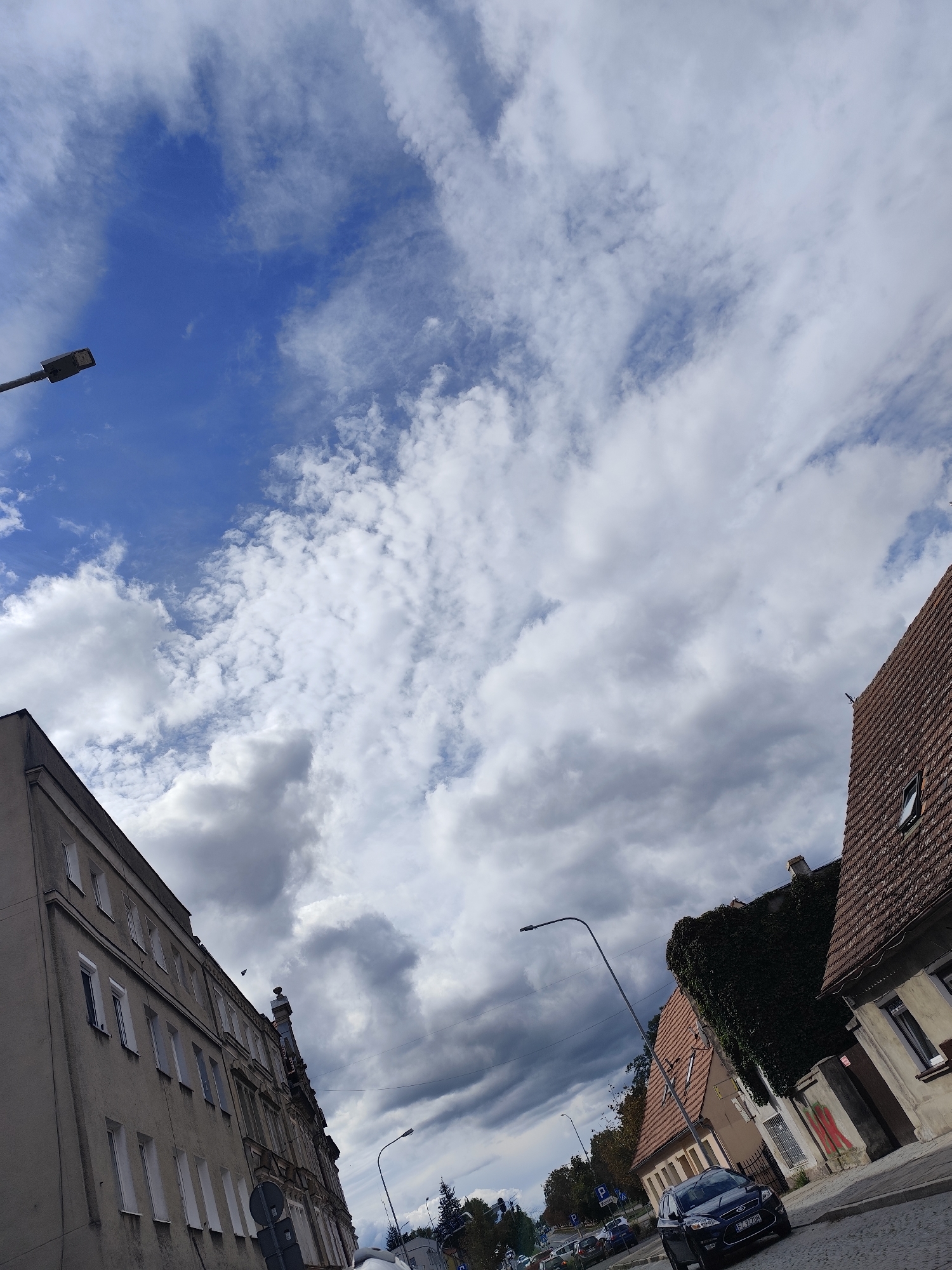 The image depicts a street scene in Zielona Góra, Poland, under a sky filled with dramatic clouds. A dark-colored Ford Mondeo, with license plate SZ 7210W, is parked on the cobblestone street, angled slightly towards the viewer. To the right of the car, a building with a terracotta-tiled roof and ivy-covered walls is visible. Across the street, a taller, older building with multiple windows dominates the left side of the frame. A streetlamp with a dark head stands near the Ford Mondeo, and another streetlamp is visible further down the street. The overall impression is a typical urban street with a mix of older and more modern architecture, set against a dynamic sky.
