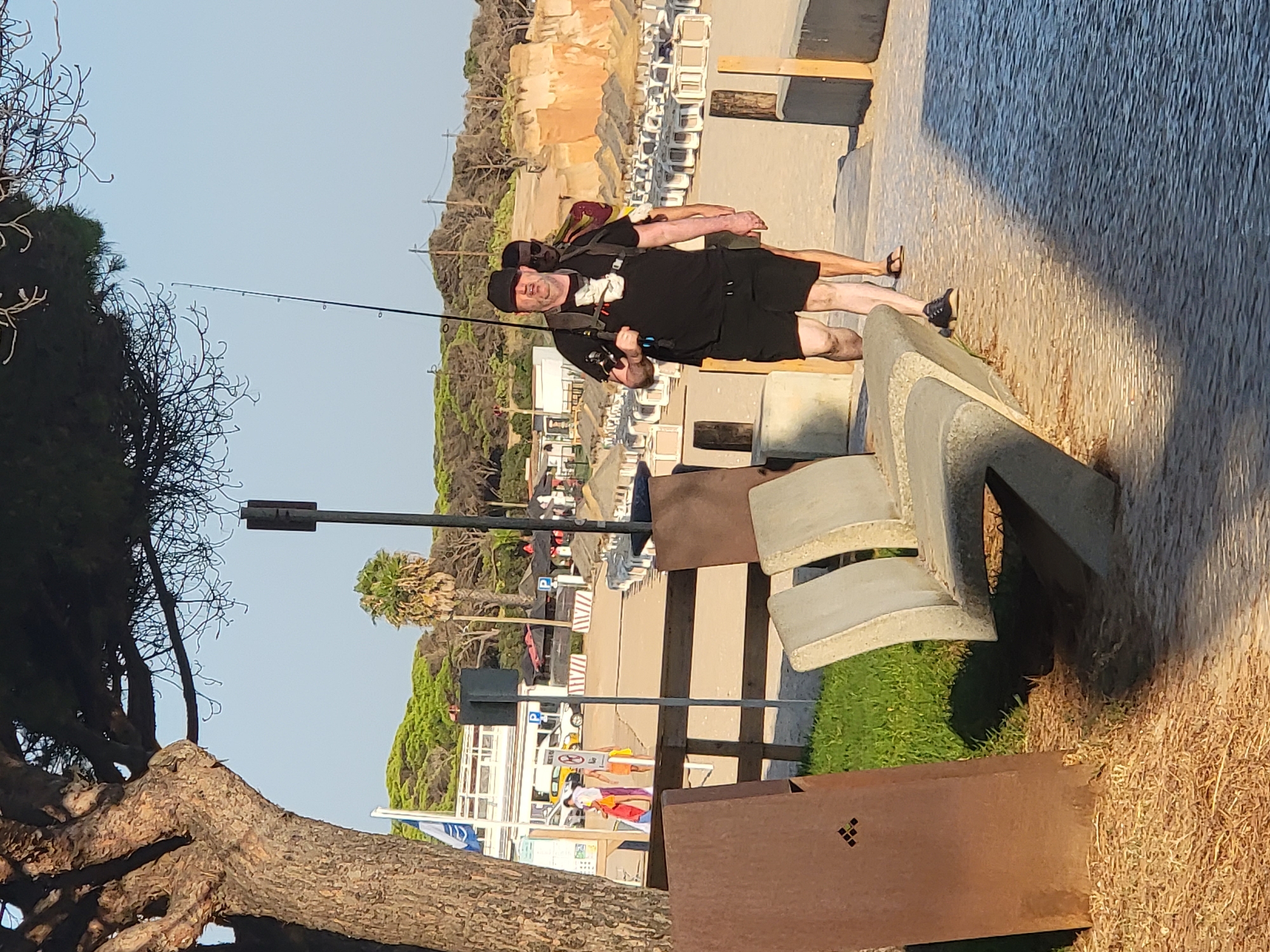 The image shows two people on a paved walkway near a beach. One person in the foreground is wearing a black t-shirt, black shorts, a black cap, and sunglasses. They are holding a fishing rod. Behind them, another person is visible, also wearing dark clothing. The background features beach chairs, sand, vegetation, and buildings. The sky is clear and blue, suggesting it is daytime. The scene appears to be a beach or promenade area. The image is oriented vertically, making it difficult to determine the exact time of day, but the lighting suggests it might be late afternoon or early evening. There is no visible text in the image.