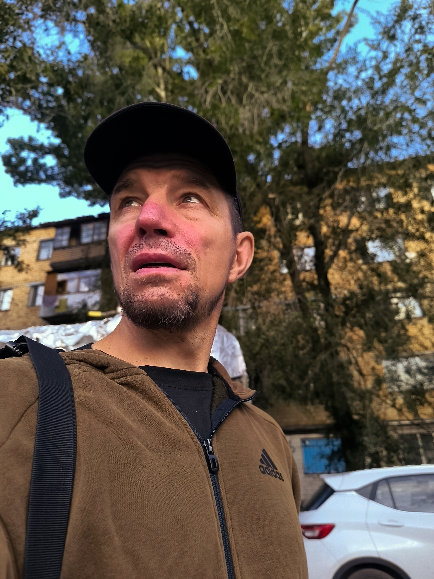 An outdoor, eye-level, medium shot captures a man with tanned skin and a light beard, looking upward and to his right. He wears a black baseball cap, a brown zippered hoodie with the Adidas logo on the left chest, and a black t-shirt underneath. A dark strap is visible across his shoulder. Behind him, a blurred background shows the branches of a tree against a bright blue sky, a yellow apartment building, and a white car. The scene is situated in the city of Qarağandı city administration, Kazakhstan.
