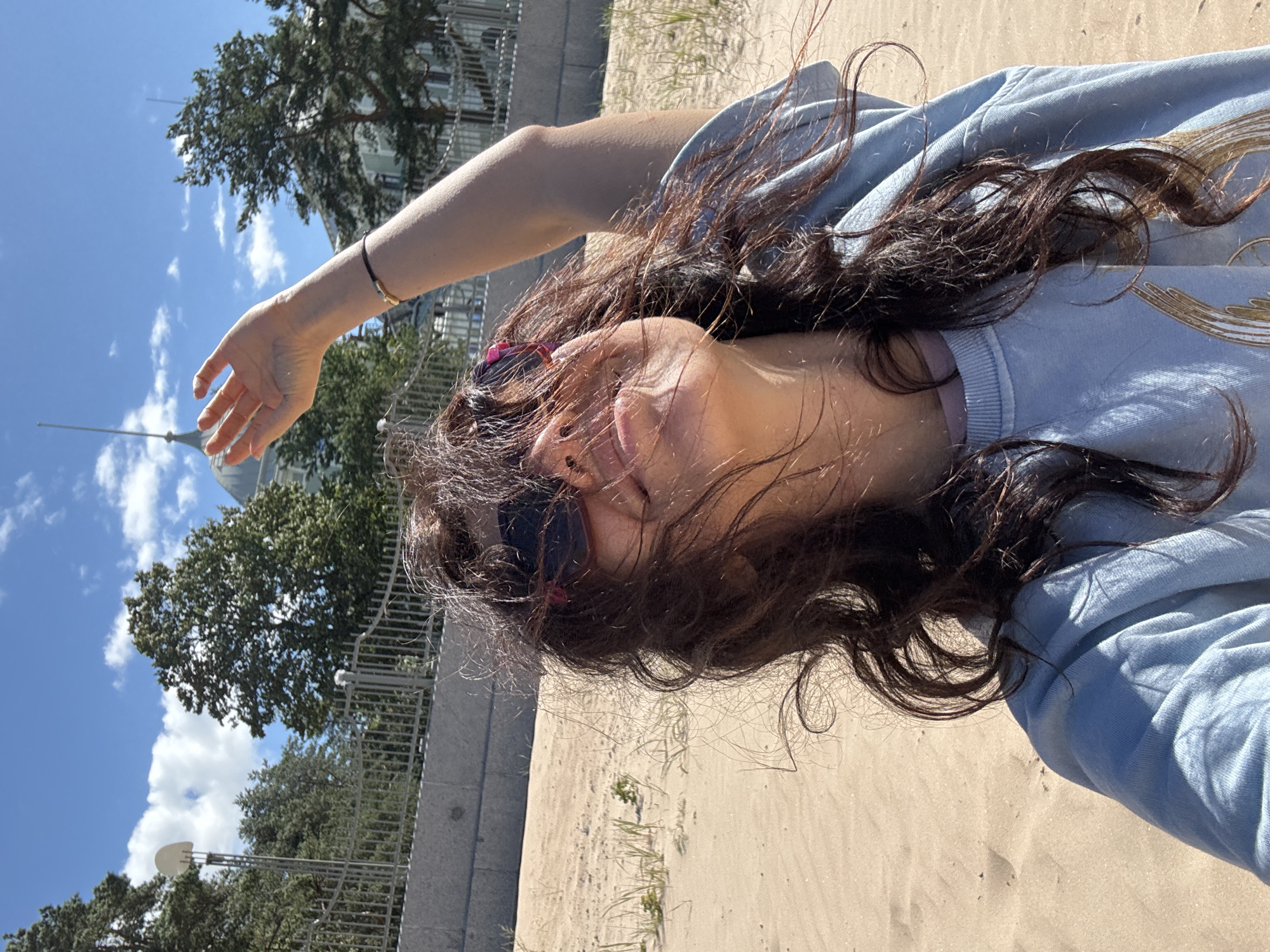 A woman with dark, windswept hair and sunglasses takes a selfie on a sandy beach. She is wearing a light blue sweatshirt. Her right arm is raised, and her hand is extended towards the sky. In the background, there is a fence, trees, a building with a dome and spire, and a bright blue sky with scattered clouds. The location is identified as being in the city of Unknown, Unknown.