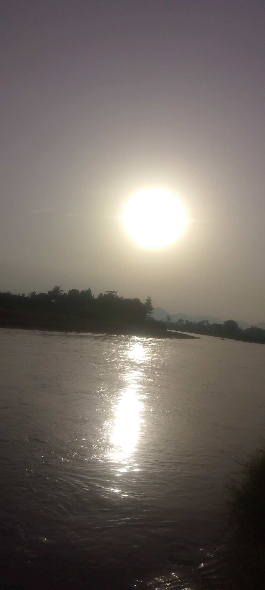 The image depicts a serene natural landscape, likely a riverbank, under a hazy sky with a prominent, bright sun. The sun, positioned high and slightly to the left, casts a strong reflection on the water, creating a shimmering pathway of light that stretches towards the foreground. The river itself appears wide and calm, with gentle ripples disturbing the surface.
Along the far bank, a dense line of silhouetted trees and foliage forms a dark, irregular horizon. These trees suggest a natural, undeveloped area. In the distance, beyond the trees, faint outlines of rolling hills or mountains can be discerned, adding depth to the scene. The overall lighting suggests either early morning or late afternoon, with the sun's rays diffused by atmospheric conditions.
There are no visible people, buildings, or man-made structures in the image, indicating a remote or natural setting. The focus is on the interplay of light and water, and the natural contours of the land.
The location is specified as Jalingo, Nigeria. Given the natural environment, this scene could be along one of the rivers or bodies of water that exist in or around the city. Jalingo is known for its proximity to mountainous regions and is situated in a savanna environment, which aligns with the depicted vegetation and landscape. The presence of a river with reflections of the sun is consistent with the geography of the Taraba State capital.