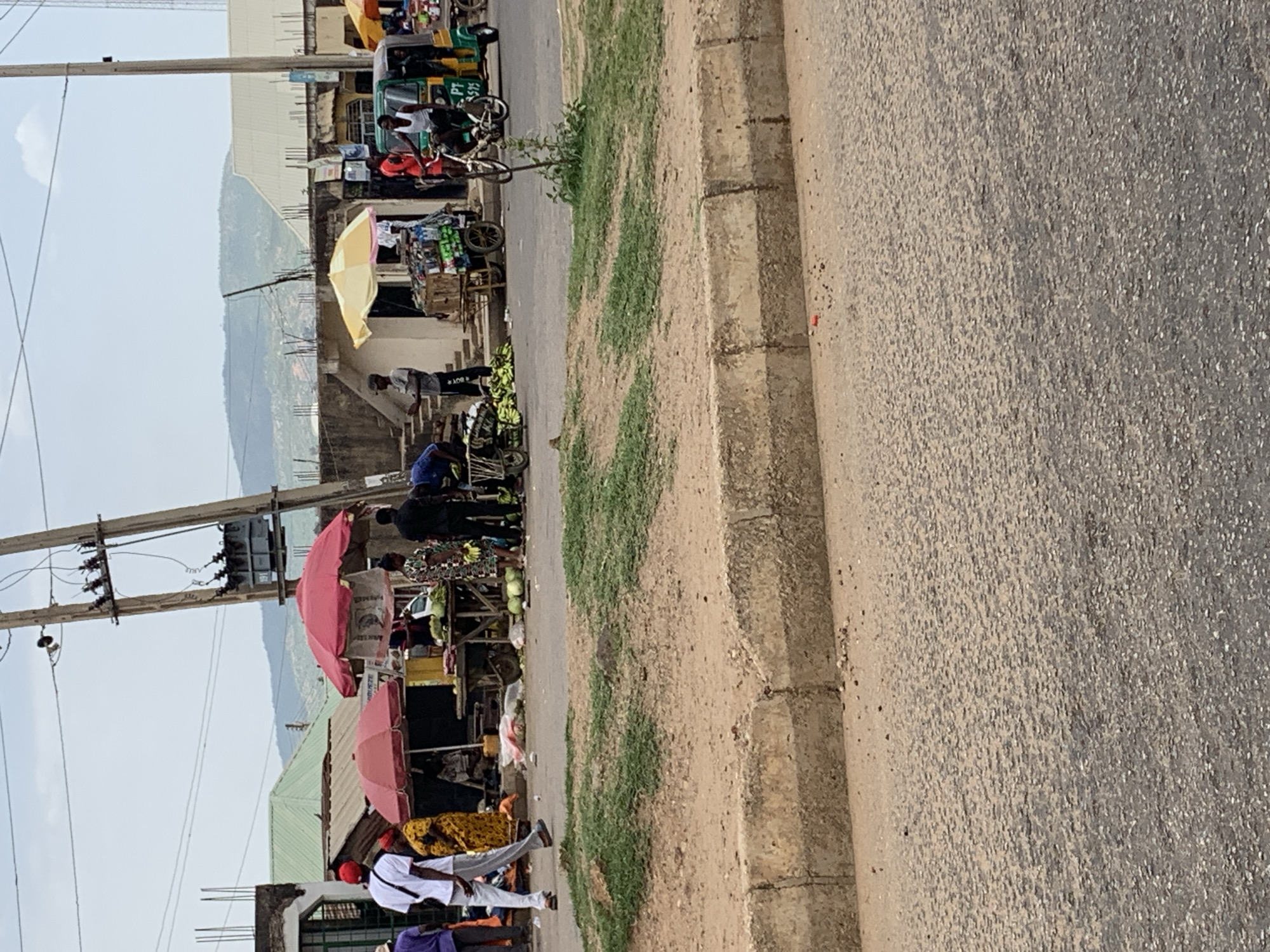 The image depicts a street scene in Rafin Par, Nigeria, with a focus on a roadside market. Several people are visible, some standing and appearing to shop, others behind makeshift stalls covered by umbrellas, likely selling goods. There are vendors displaying their wares. The environment is urban, with buildings and a distant hillside visible. Utility poles with electrical transformers are present. The street is paved, and a curb separates the road from the market area. A motorized tricycle taxi is parked near one of the stalls.