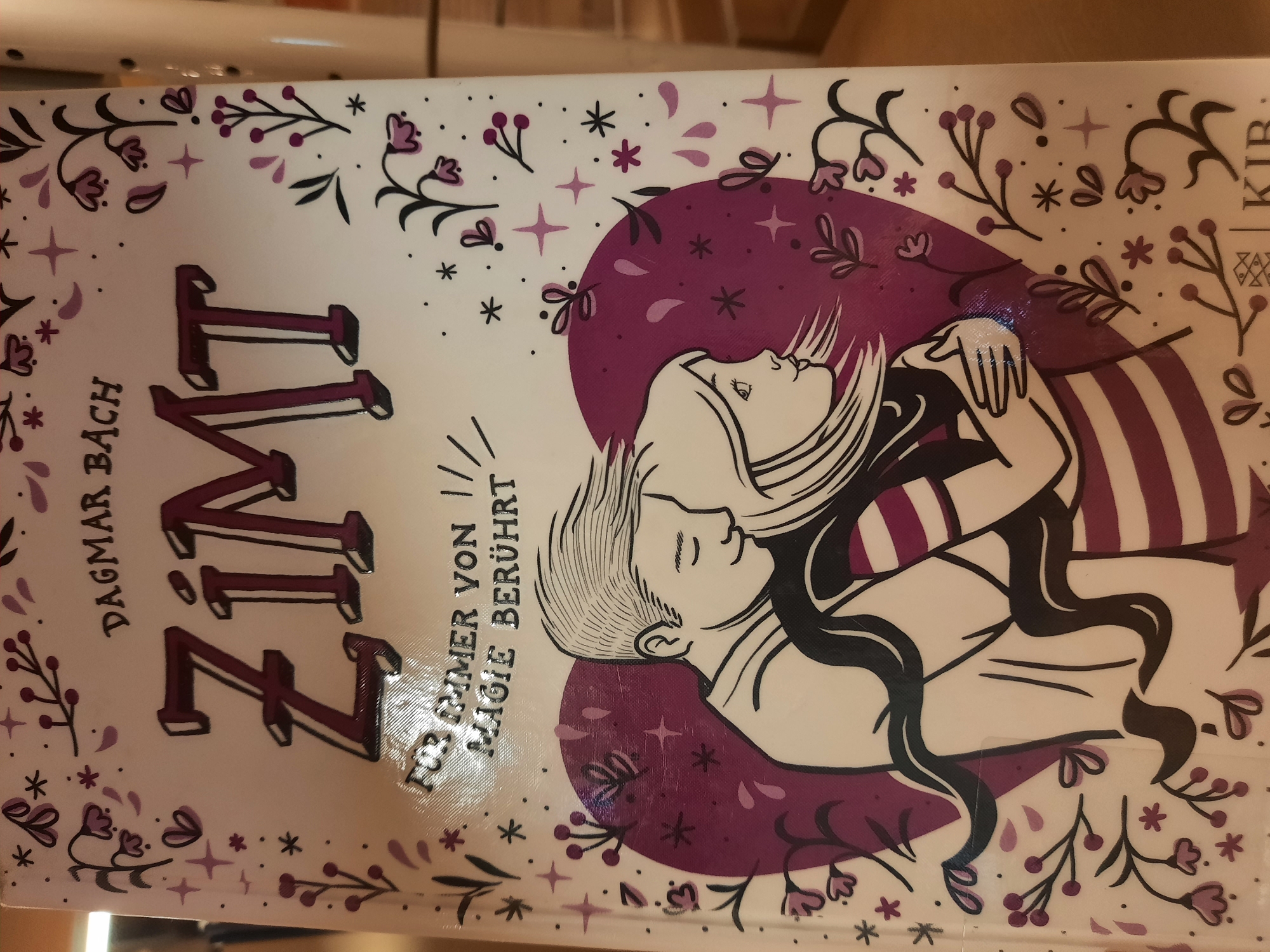 A close-up, eye-level, indoor shot showcases the cover of a book titled "Zimt" by Dagmar Bach. The book cover features a stylized illustration in shades of purple and white against a white background, with decorative floral and star motifs bordering the edges. The central illustration depicts a young couple embracing tightly, with the man's arms wrapped around the woman. The woman's hair is long and flowing, and she is wearing a striped shirt. The text on the cover reads "DAGMAR BACH" in the upper center and "FÜR IMMER VON MAGIE BERÜHRT" below the title "ZIMT". The image's lighting suggests it was taken in a well-lit indoor environment. The book appears to be displayed on a shelf or surface, with a blurred wooden background visible to the right. This book cover is a product of KJB. No specific location in the city of Rosenheim, Germany, is depicted.