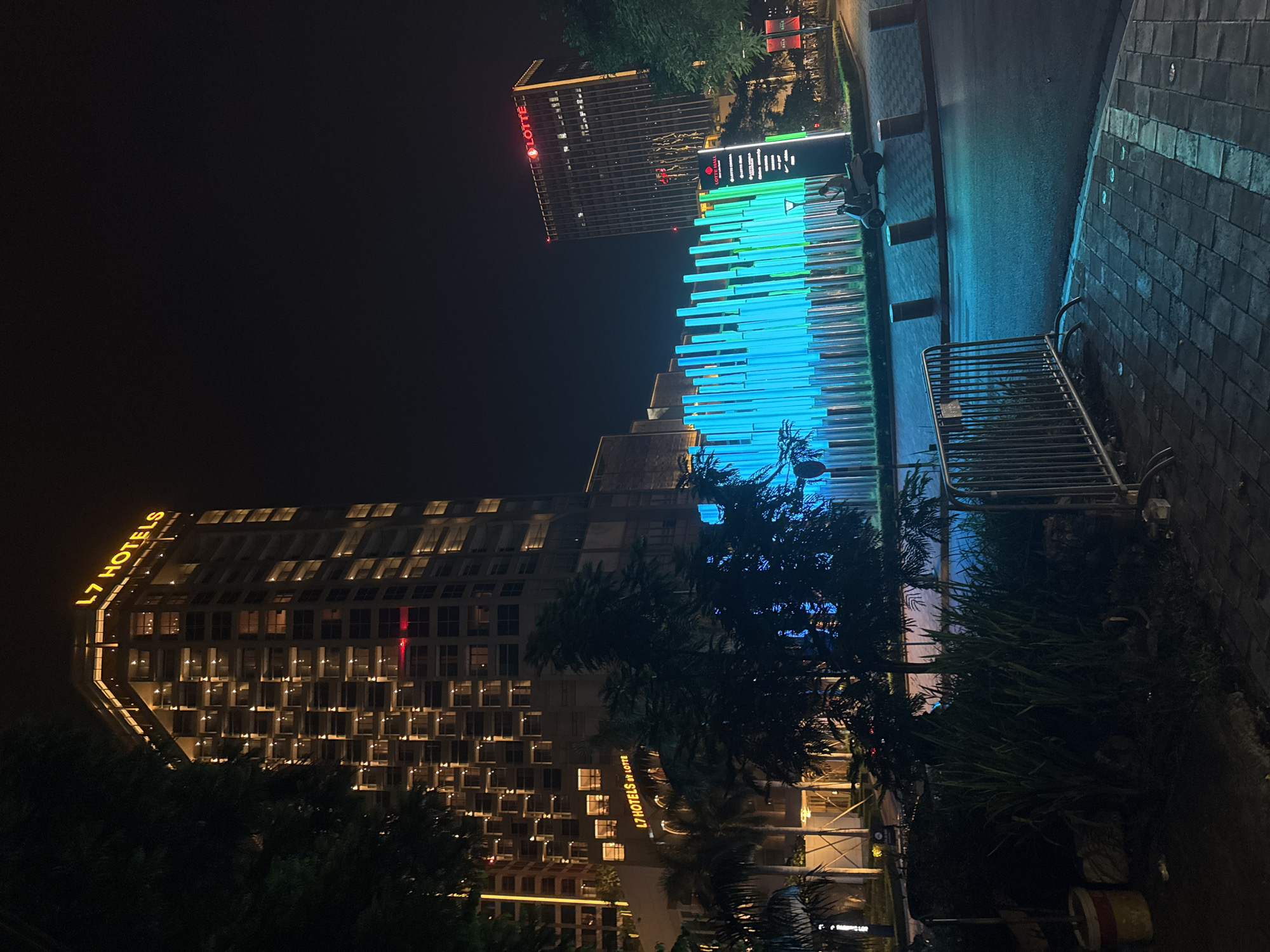 The image captures a nighttime view of a modern urban landscape in Hanoi, Vietnam. Dominating the left side is a tall, illuminated building with the "L7 HOTELS by LOTTE" signage visible. The building features numerous windows, many of which are lit, creating a grid-like pattern against the dark sky. In the background to the right, another tall building displays the "LOTTE" logo in red neon. In the mid-ground, a striking installation of vertical, colored lights, predominantly blue and green, creates a vibrant focal point. A person is riding a scooter past this installation, and a few bollards line the street. The foreground shows a paved walkway with cobblestone patterns, a metal barrier, and some vegetation. The scene suggests a well-developed commercial or entertainment district within the city.