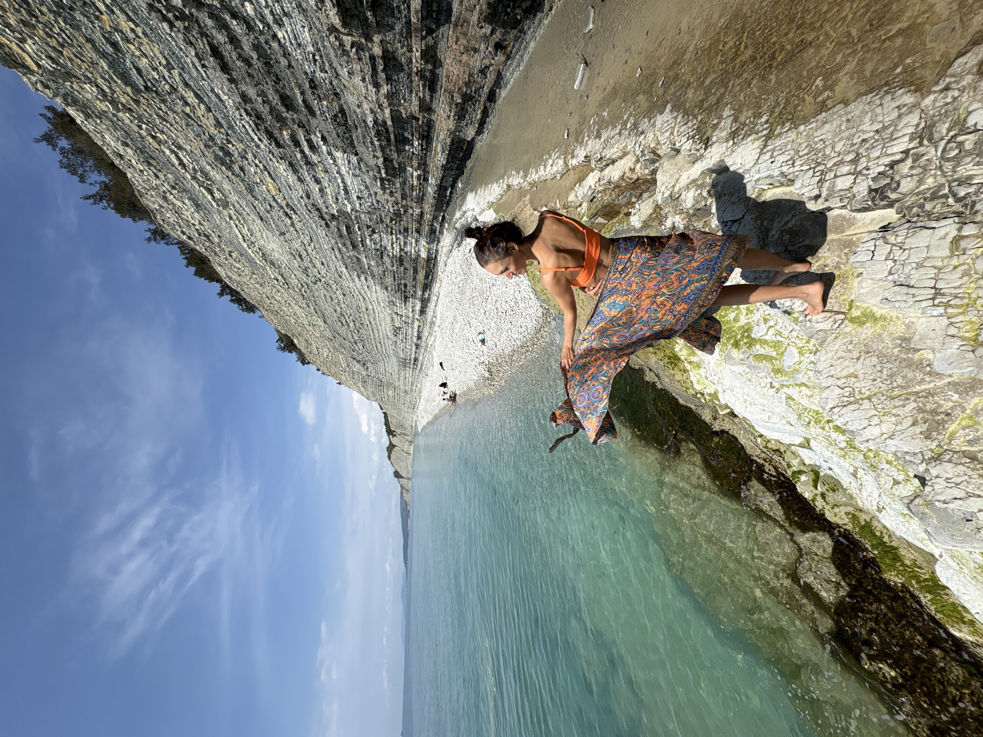 This image, set in Gelendzhik, Russia, captures a scenic coastal view on a bright, sunny day under a clear blue sky with sparse, wispy clouds.
The central subject is a woman standing barefoot on a rocky shore next to the clear, turquoise-colored sea. She has dark hair styled in a bun and is wearing an orange bikini top paired with a flowing, vibrantly patterned maxi skirt featuring intricate designs in shades of blue, orange, and purple. She is bending forward, looking down intently at the water's edge or the rocks immediately in front of her, suggesting a moment of observation or interaction with her natural surroundings.
The backdrop is dominated by an impressive, towering cliff face, characterized by distinct horizontal layers of greyish rock, possibly shale or limestone. This dramatic geological formation rises steeply from the narrow, pebbly beach, with sparse green vegetation visible along its upper ridge. The sea water is remarkably clear near the shore, revealing a rocky bottom covered in patches of green algae, while further out, the water transitions to a deeper blue. In the distance, along the curve of the coastline, faint figures suggest the presence of other people on the beach. Strong sunlight casts sharp shadows, highlighting the textures of the rocks and the woman's form. There is no visible text in the image.