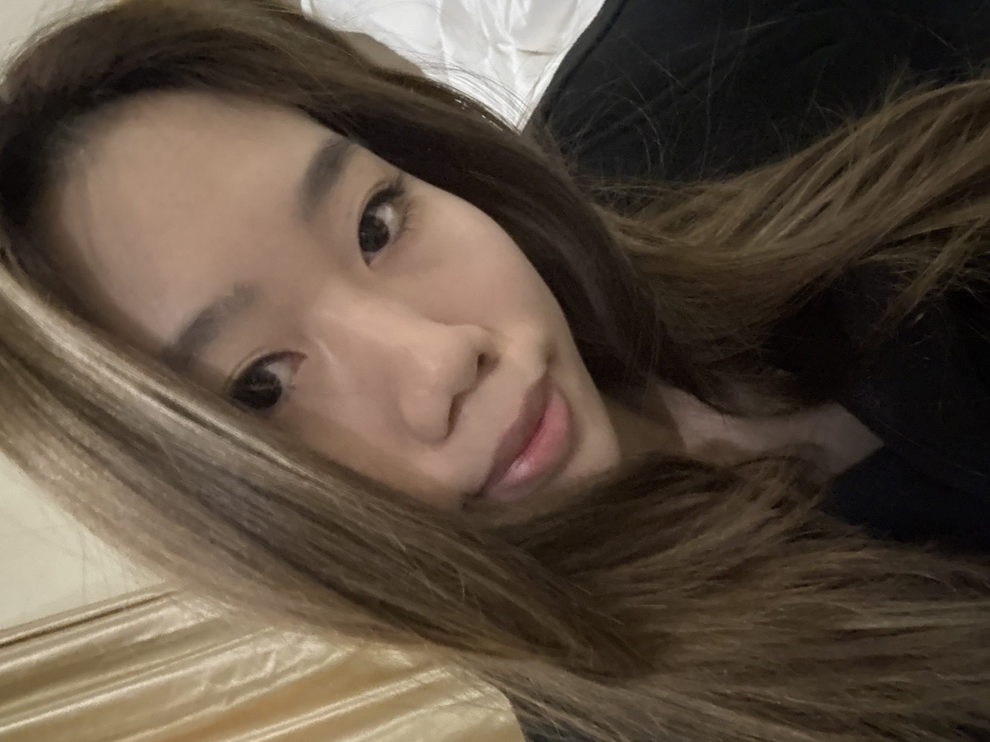 A close-up, selfie-style photograph depicts an Asian woman with long brown hair. Her hair is styled to fall across her face and shoulders. Her facial features are clearly visible, including her eyes, nose, and lips. She appears to be lying down, possibly in bed, as indicated by the golden satin-like material visible at the lower-left edge of the frame. The background consists of dark clothing, possibly a shirt or jacket. The image does not contain sufficient information to determine a specific location in Dunhuang City, China.