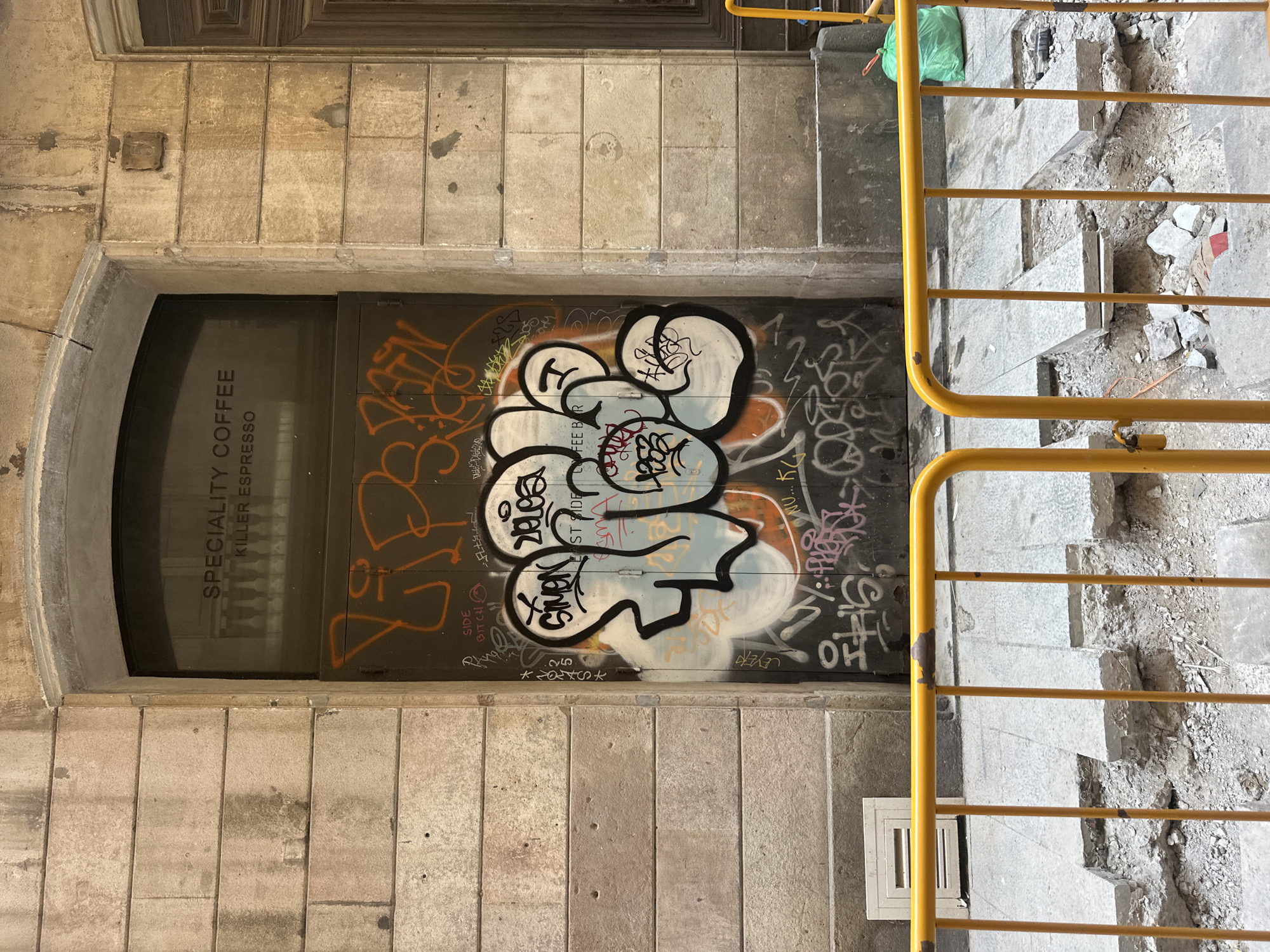 This is a vertical, eye-level shot of an alleyway in Barcelona, Spain. The scene is characterized by urban decay and graffiti. On the left, a weathered stone building features a dark-tinted glass door with vertical white text reading "SPECIALITY COFFEE KILLER ESPRESSO." The door and the adjacent wall are extensively covered in graffiti, with bold, colorful tags in orange, black, and white. The main graffiti on the door appears to be a cartoonish character or abstract design.
To the right of the door, a set of stone stairs is under construction or renovation, with rubble and broken concrete scattered around. A bright yellow metal railing lines the stairs, partially obscuring the view of the steps. A bright green plastic bag is visible near the top of the stairs. The overall impression is one of a gritty, urban environment, possibly in a less-trafficked area, with a contrast between the commercial signage and the pervasive street art and ongoing construction. The lighting suggests it is daytime. There is no visible text on the yellow railing or the green bag.