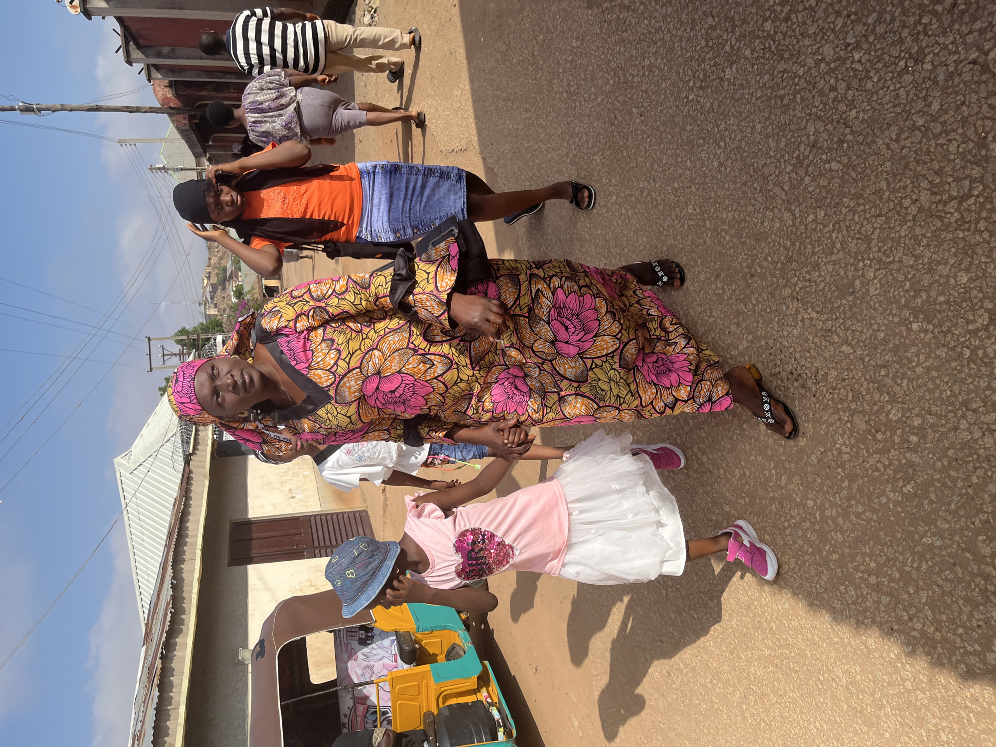 On a street in Jos, Nigeria, a woman in a colorful dress and headwrap holds the hand of a young girl in a pink shirt and white skirt. The girl wears a blue hat and pink sneakers. They stand next to a three-wheeled vehicle known as a Keke Napep. Behind them, two other figures are walking, one in an orange shirt and the other in a striped shirt. The ground is paved, and buildings are visible in the background.