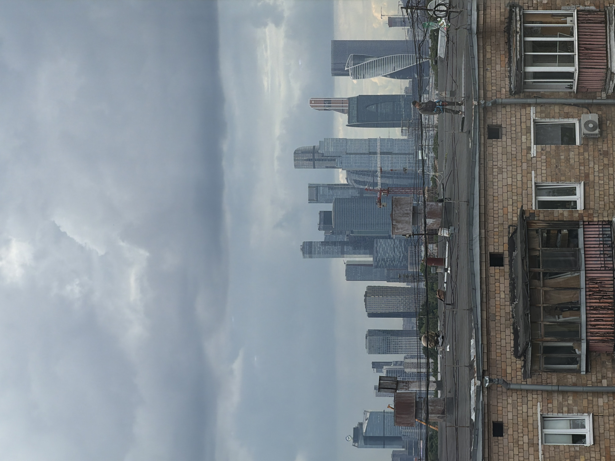 This is a photograph of a cityscape with a focus on an older brick building in the foreground and modern skyscrapers in the background. A person is visible on a rooftop, appearing to be engaged in some kind of work at height, possibly window cleaning or maintenance, as they are wearing a harness and have equipment attached. The person is looking away from the camera.
The scene is set against a cloudy sky, suggesting it might be an overcast day. The time of day is not explicitly clear, but the lighting appears to be diffused due to the cloud cover. The juxtaposition of the aged brick building with its worn windows and the sleek, contemporary architecture of the skyscrapers is a prominent visual element. Wires and piping are also visible on the rooftop of the brick building, adding to the urban industrial feel.
The person's activity at height on the rooftop, with the vast expanse of the city in the distance, creates a sense of scale and the human element within a large urban environment. There is no visible text in the image. The overall mood is perhaps one of daily labor set against a dynamic urban backdrop.