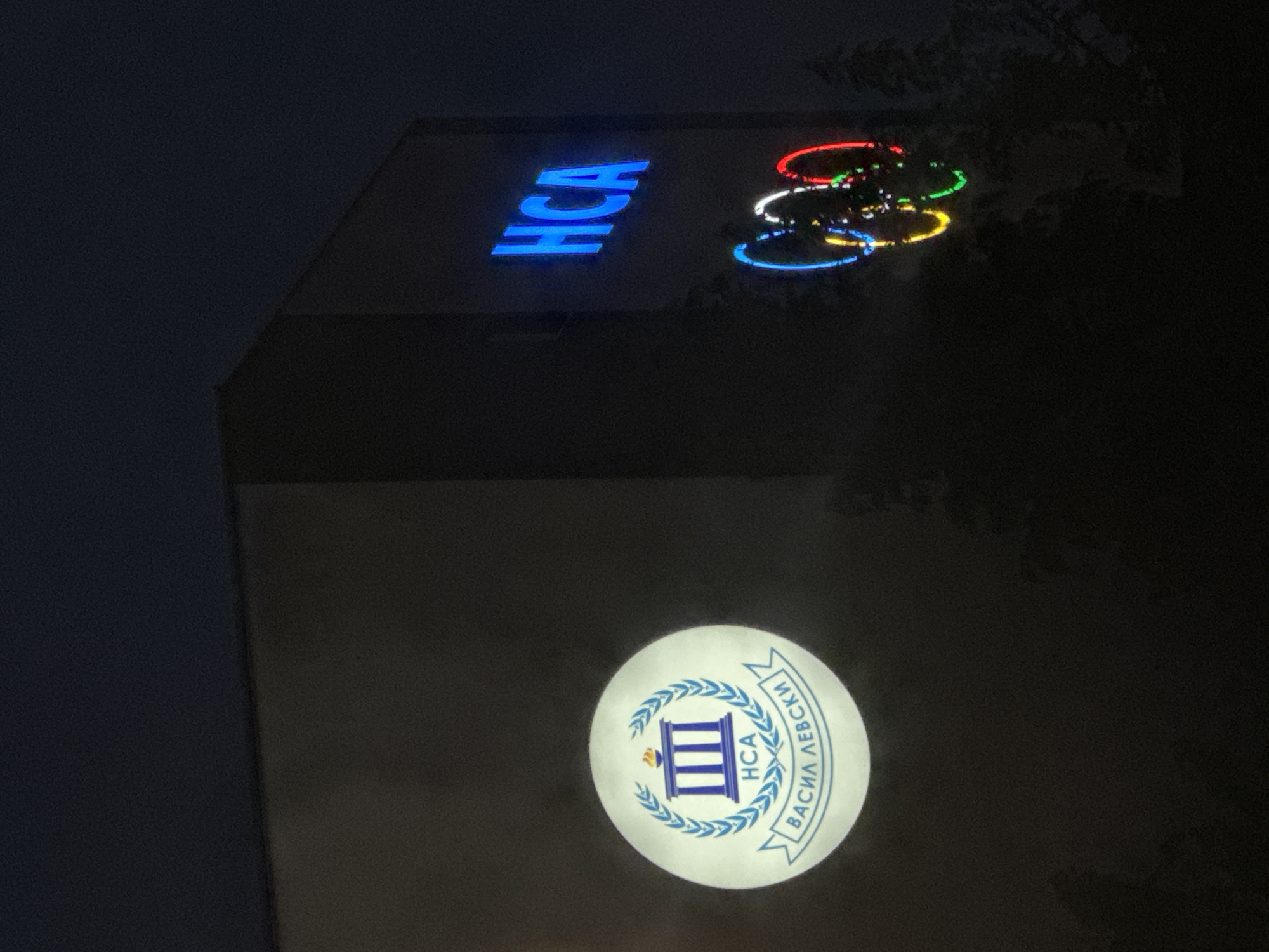 The image shows the exterior of a building at night, likely a sports facility or educational institution, in Sofia, Bulgaria. The building has a dark facade. On the left side of the building, a circular emblem is illuminated. This emblem contains a depiction of a classical building with columns, surrounded by a laurel wreath, and inscribed with the letters "HCA" and the Cyrillic text "ВАСИЛ ЛЕВСКИ". On the right side of the building, the letters "HCA" are displayed prominently in bright blue neon. Below the "HCA" sign are the Olympic rings, illuminated in their signature colors of blue, yellow, red, green, and black. The Olympic rings are slightly out of focus due to the night setting and potential movement. The background is dark, suggesting the image was taken after sunset. Silhouetted trees are visible in the foreground, partially obscuring the lower part of the building and the Olympic rings. The overall impression is of an official building decorated with its institutional emblem and the Olympic symbol, signifying a connection to sports or education, possibly within the National Sports Academy "Vasil Levski."