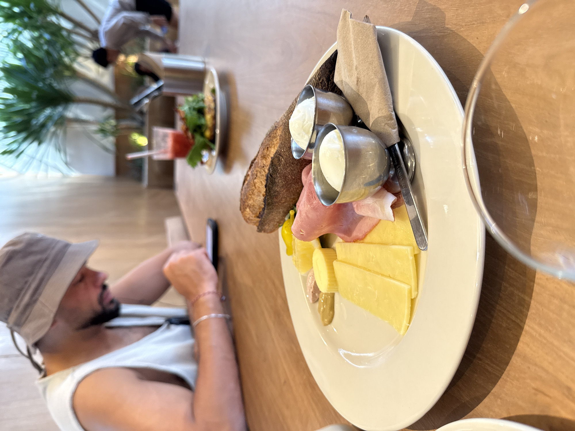 The image shows a restaurant scene, likely in North Kuta, Indonesia. A fair-skinned man wearing a bucket hat and a white tank top is reclining on a bench, looking at his mobile phone. His left arm is visible, adorned with two bracelets.
On the table in front of him is a white plate with an assortment of food. The plate contains slices of bread, two small metal containers filled with what appears to be cream, several slices of yellow cheese, some cured ham, a folded beige napkin, and a spoon.
Behind the man, another table has a salad and a drink. Further in the background, there is a person wearing a white shirt. A green plant is also visible near the window. A partially-visible glass of water is on the right-hand side of the table. The restaurant has a wooden table surface.